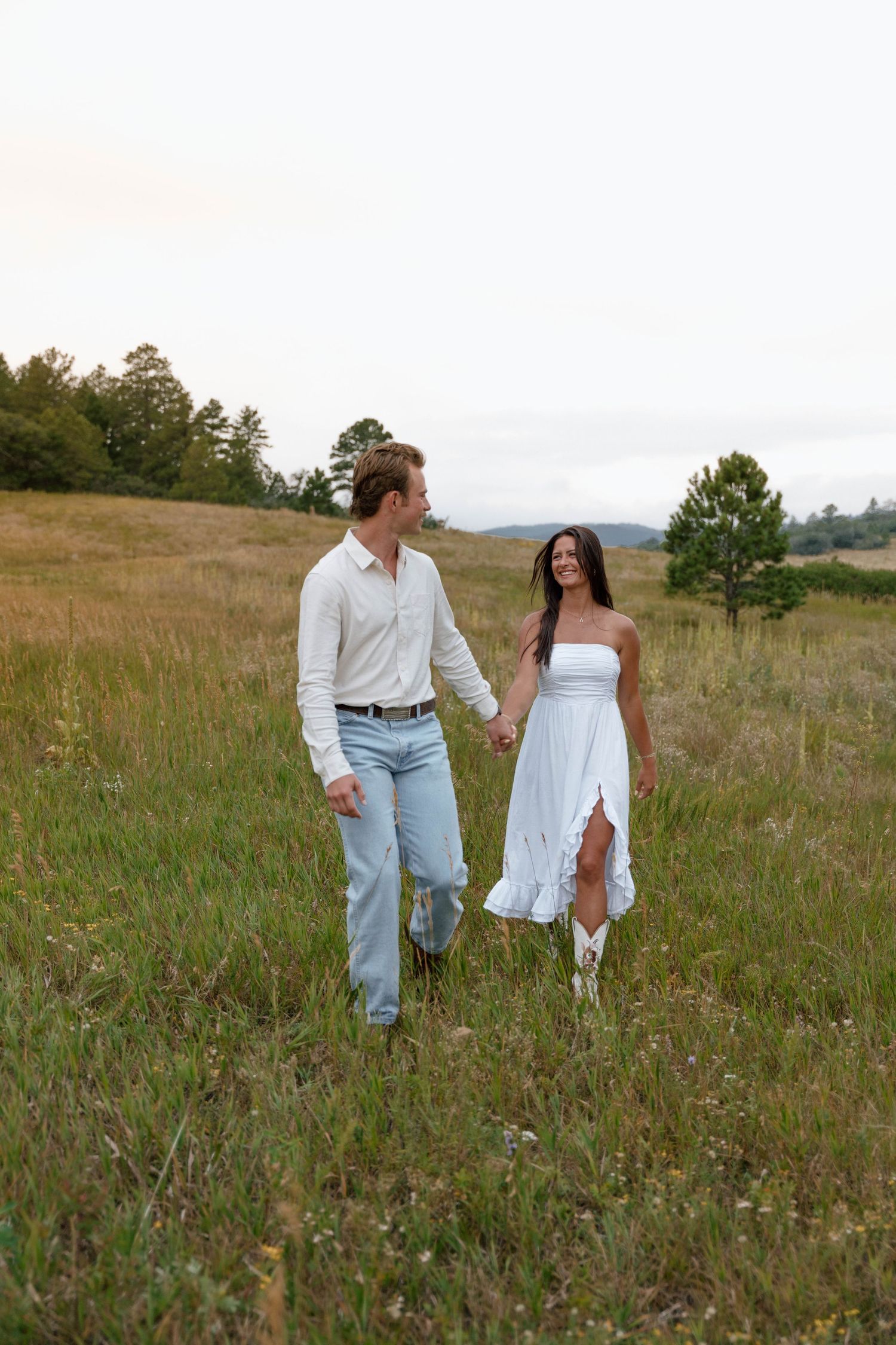 A couple walks together through a mountain meadow wearing matching white outfits at sunset.