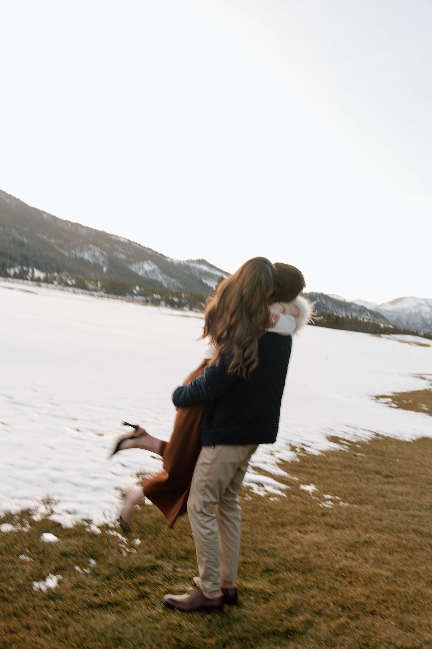 Couple embracing on snowy ground with mountains visible in the distance.