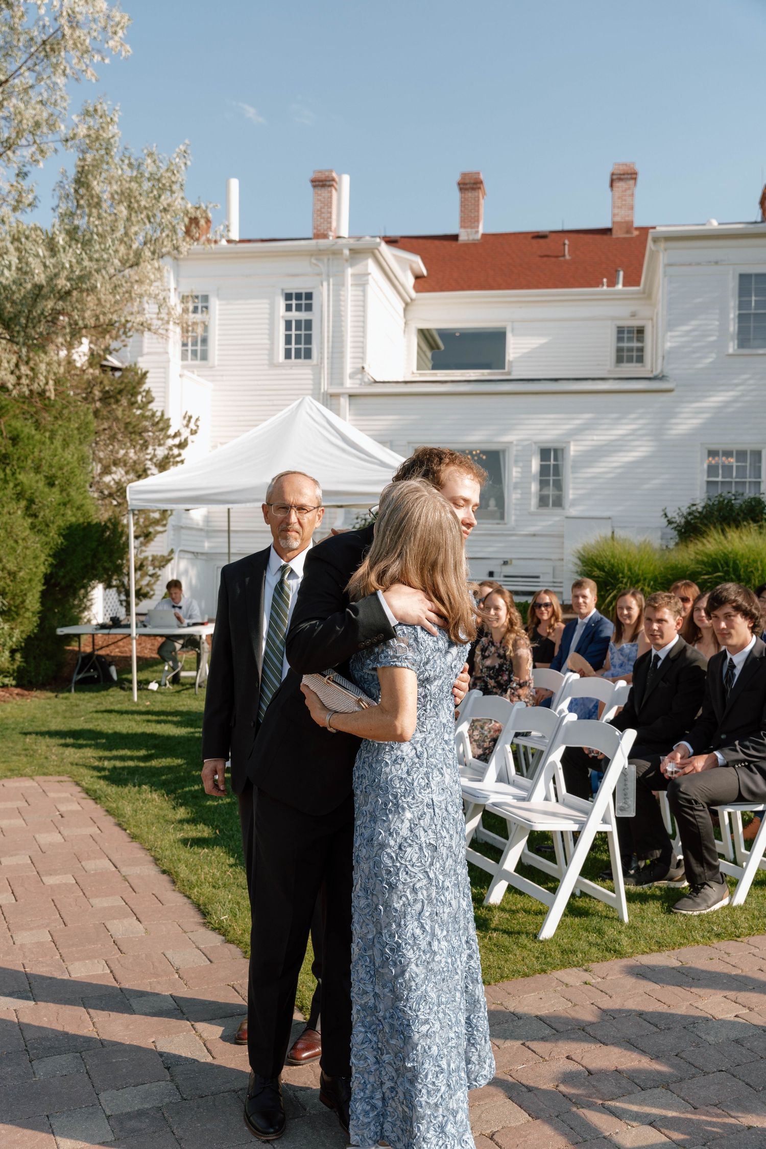 Outdoor wedding ceremony with guests seated on white chairs and brick pathway.