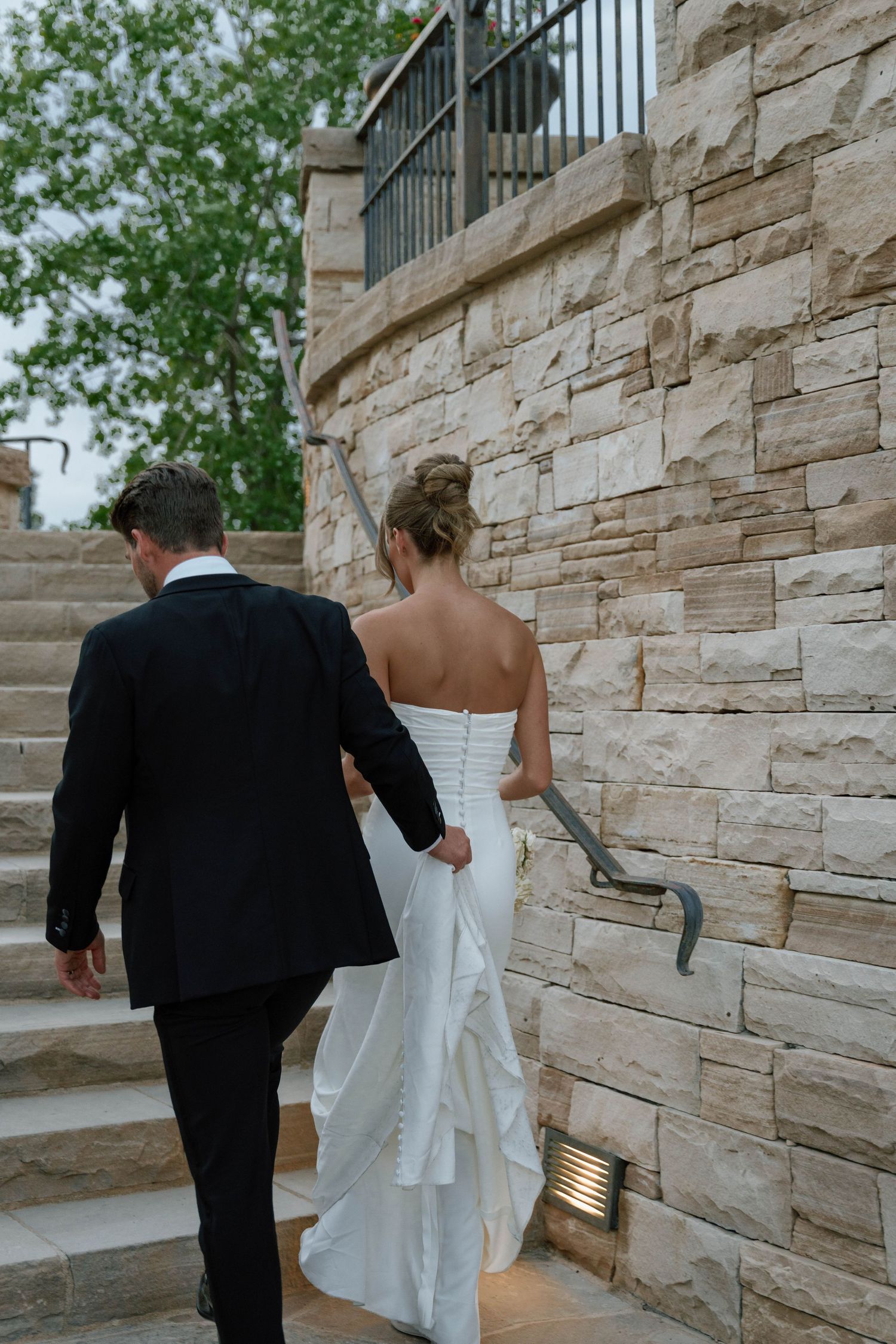 A couple walks up stone steps alongside a rustic stone wall at their wedding venue.