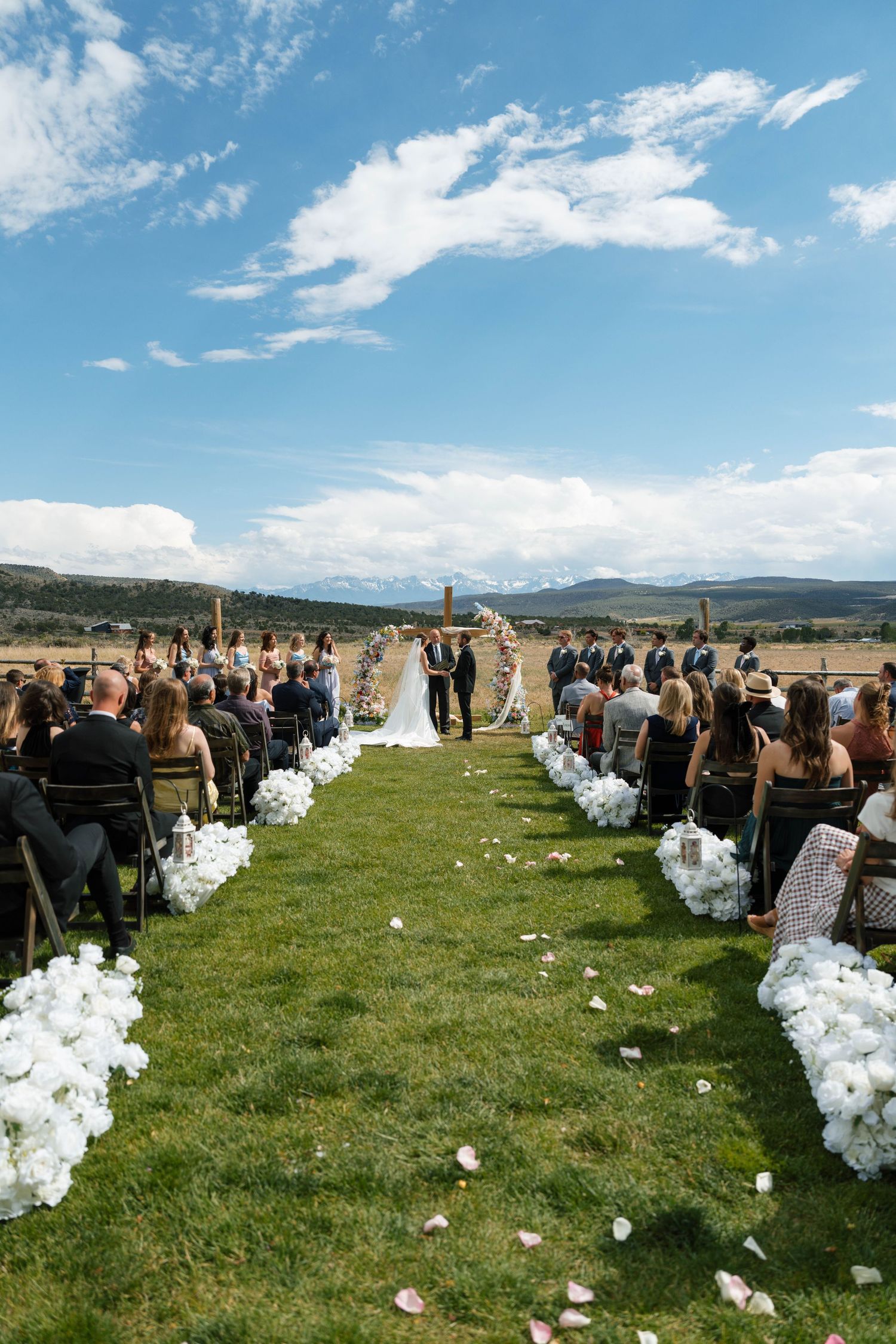 Outdoor wedding ceremony on green grass aisle lined with white floral arrangements, couple at altar with mountain views.