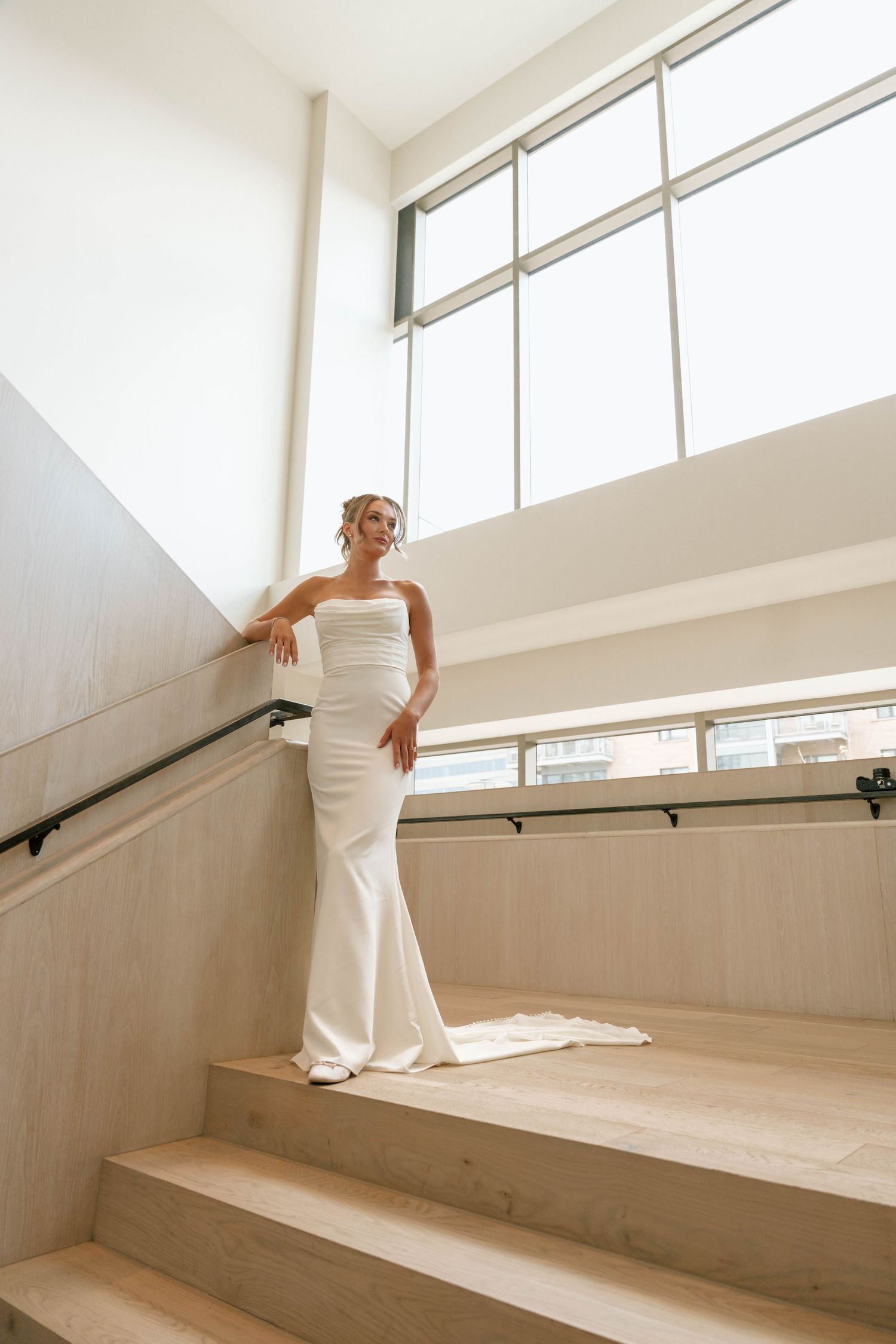 A bride stands gracefully on light wooden stairs beneath tall windows in a modern setting.