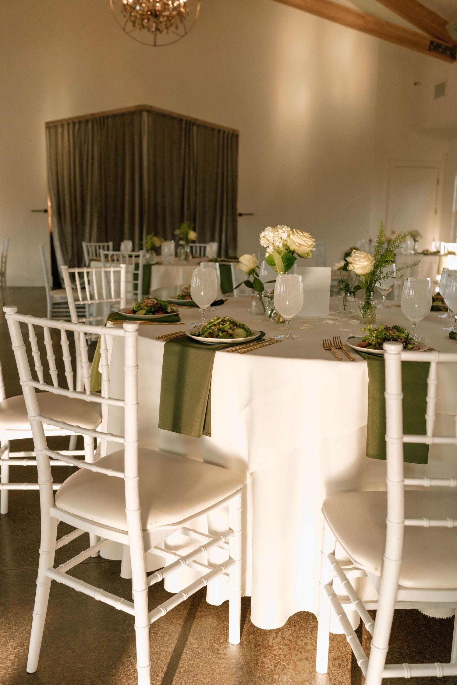 White chairs and tables elegantly arranged in a reception hall with a crystal chandelier overhead.