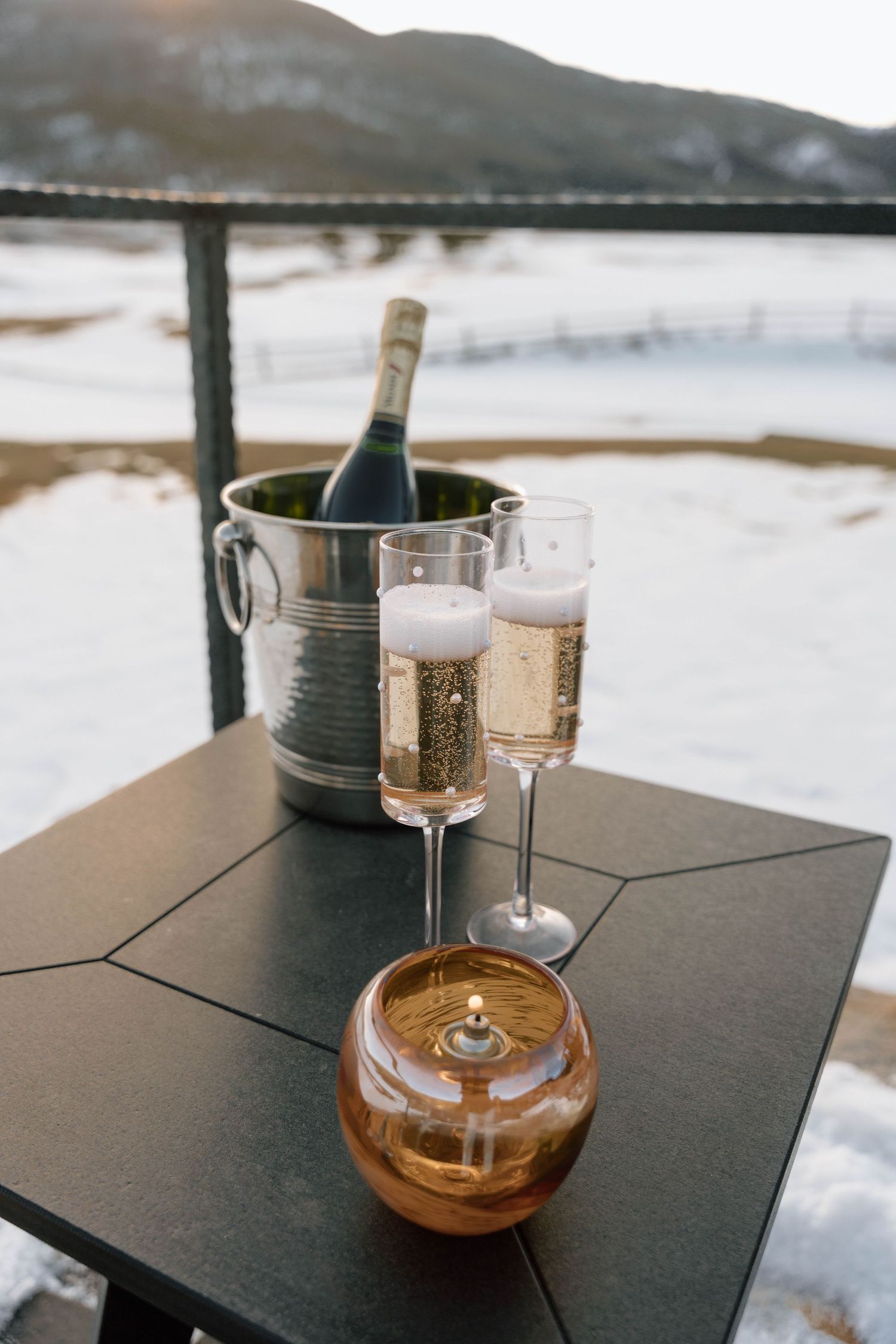 Champagne chilling in ice bucket with glasses and amber drink on outdoor table with snowy mountain view.