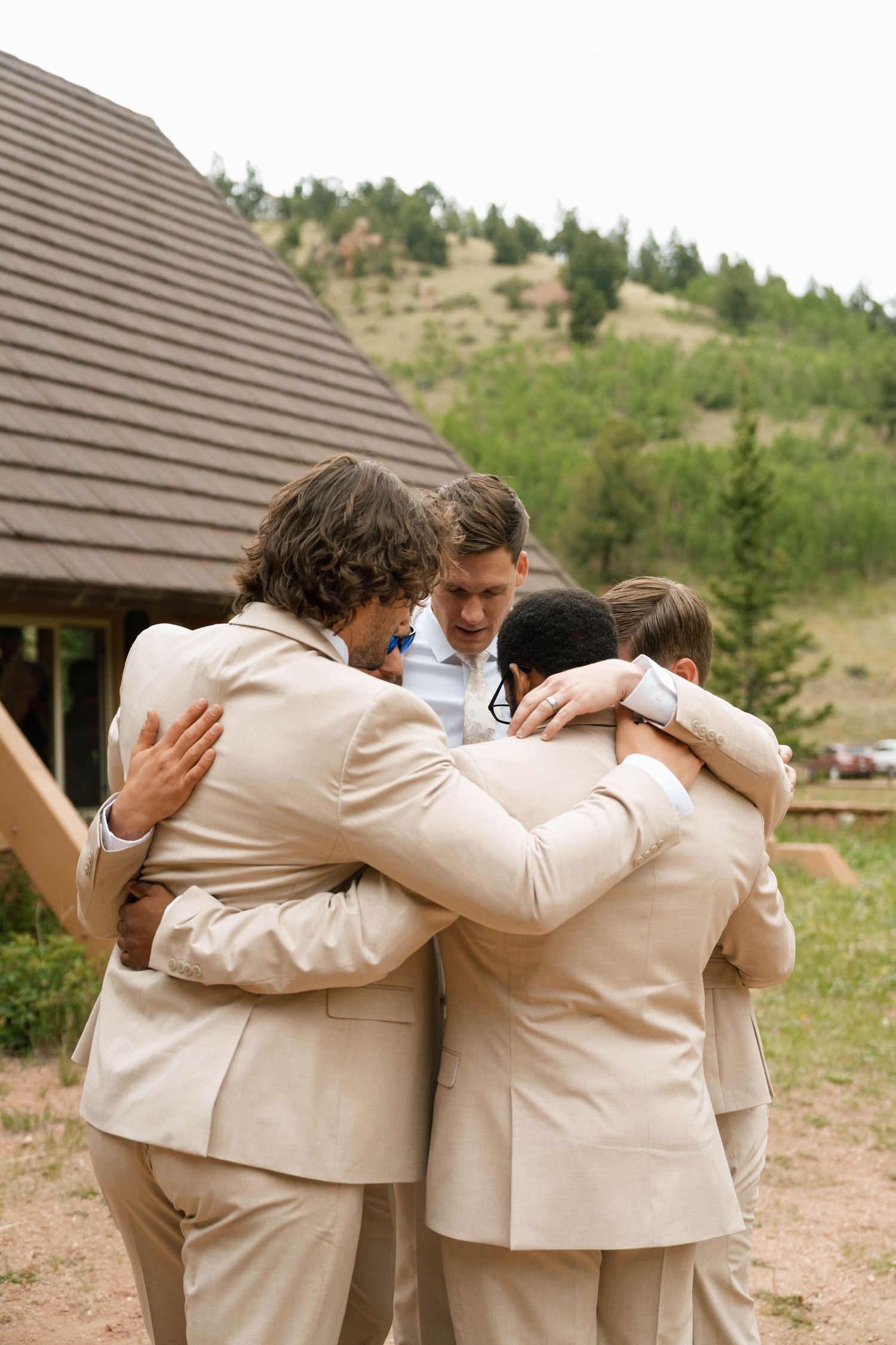 Groomsmen in matching tan suits share a group huddle outside mountain wedding venue.