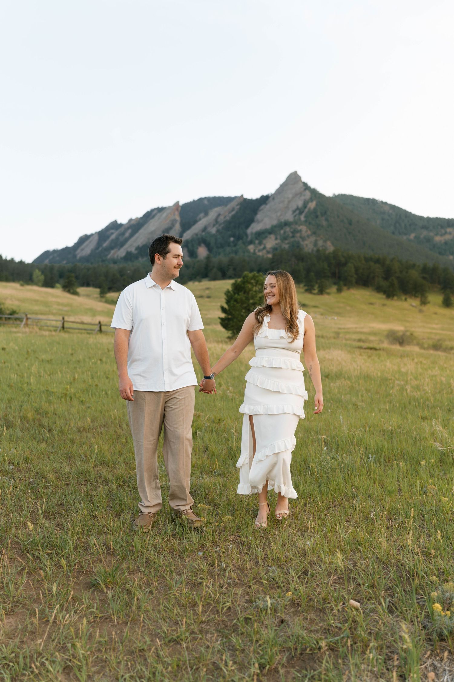 Smiling couple in white attire holding hands walking through meadow with Flatirons mountains behind them.