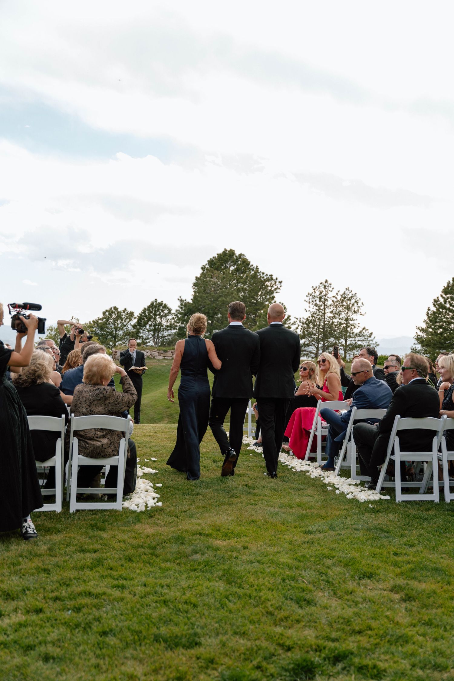Wedding guests seated in white chairs on a grassy hillside for an outdoor ceremony.
