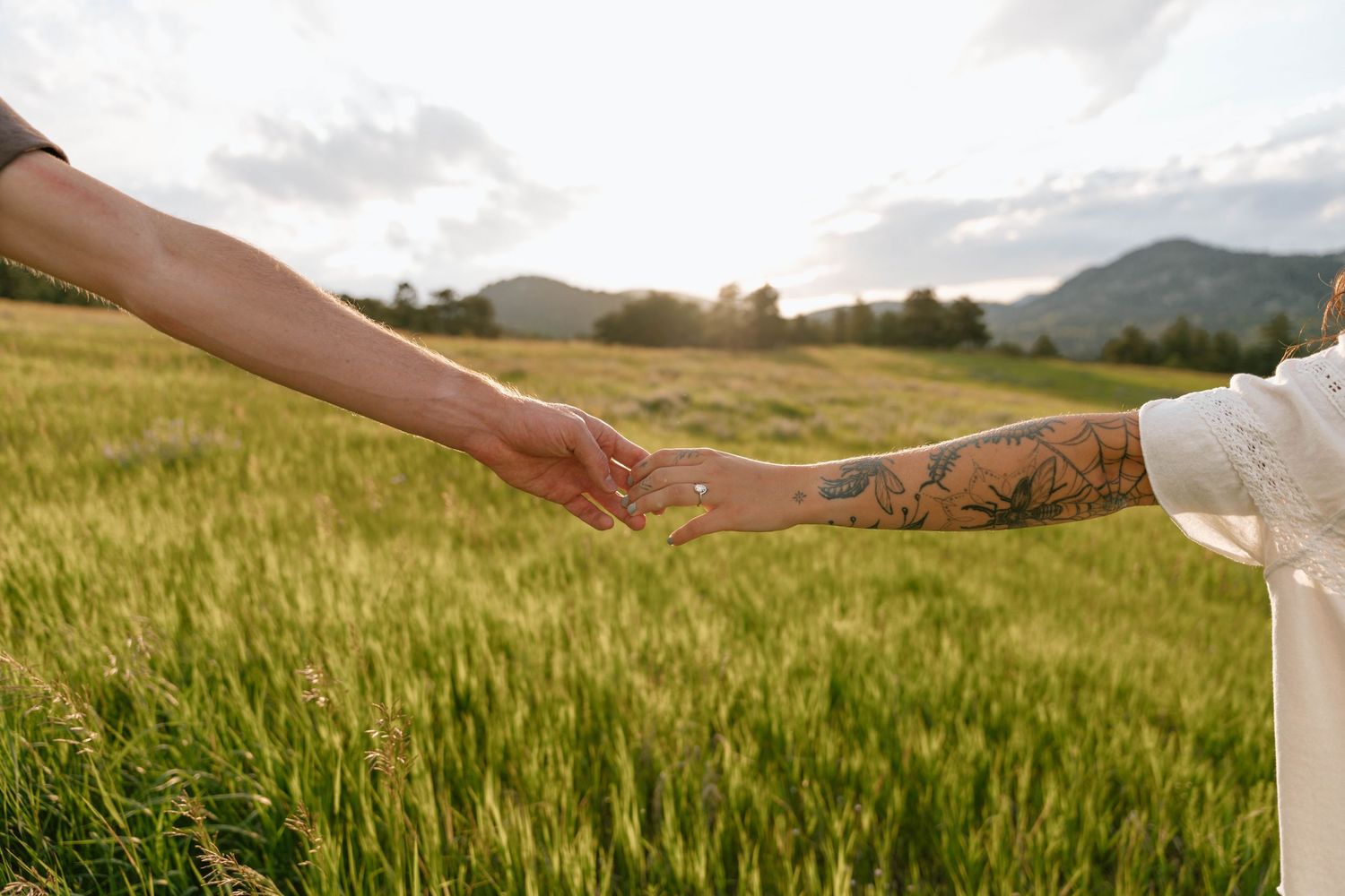Two people holding hands while walking through a scenic mountain meadow at sunset.