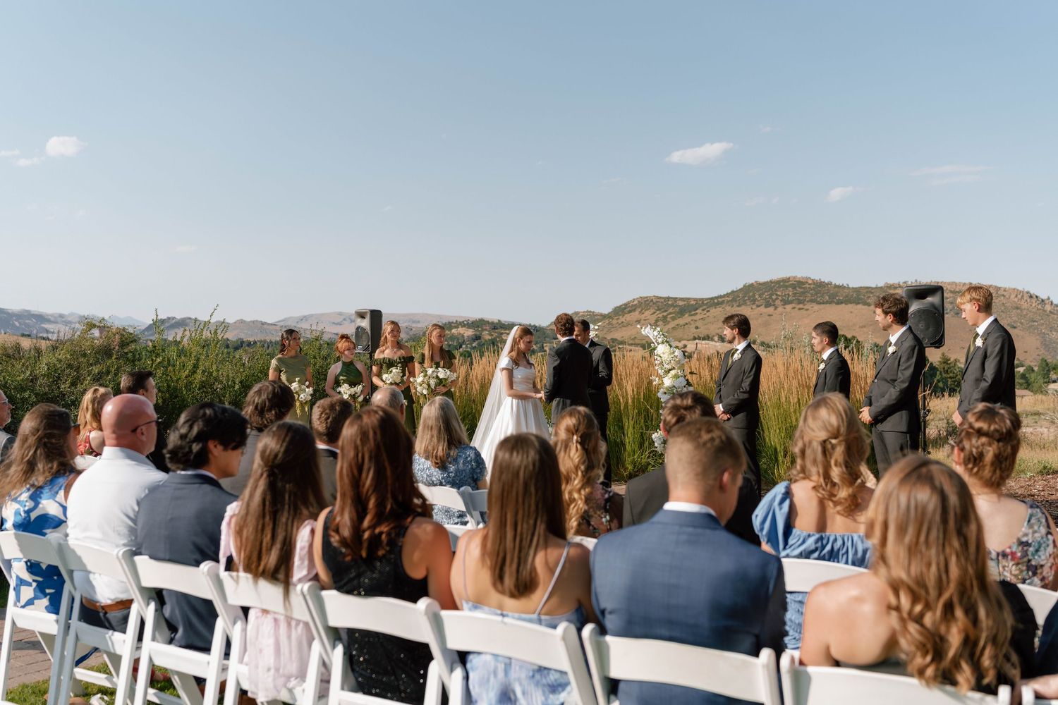 Outdoor wedding ceremony with guests seated on white chairs against a mountain backdrop.