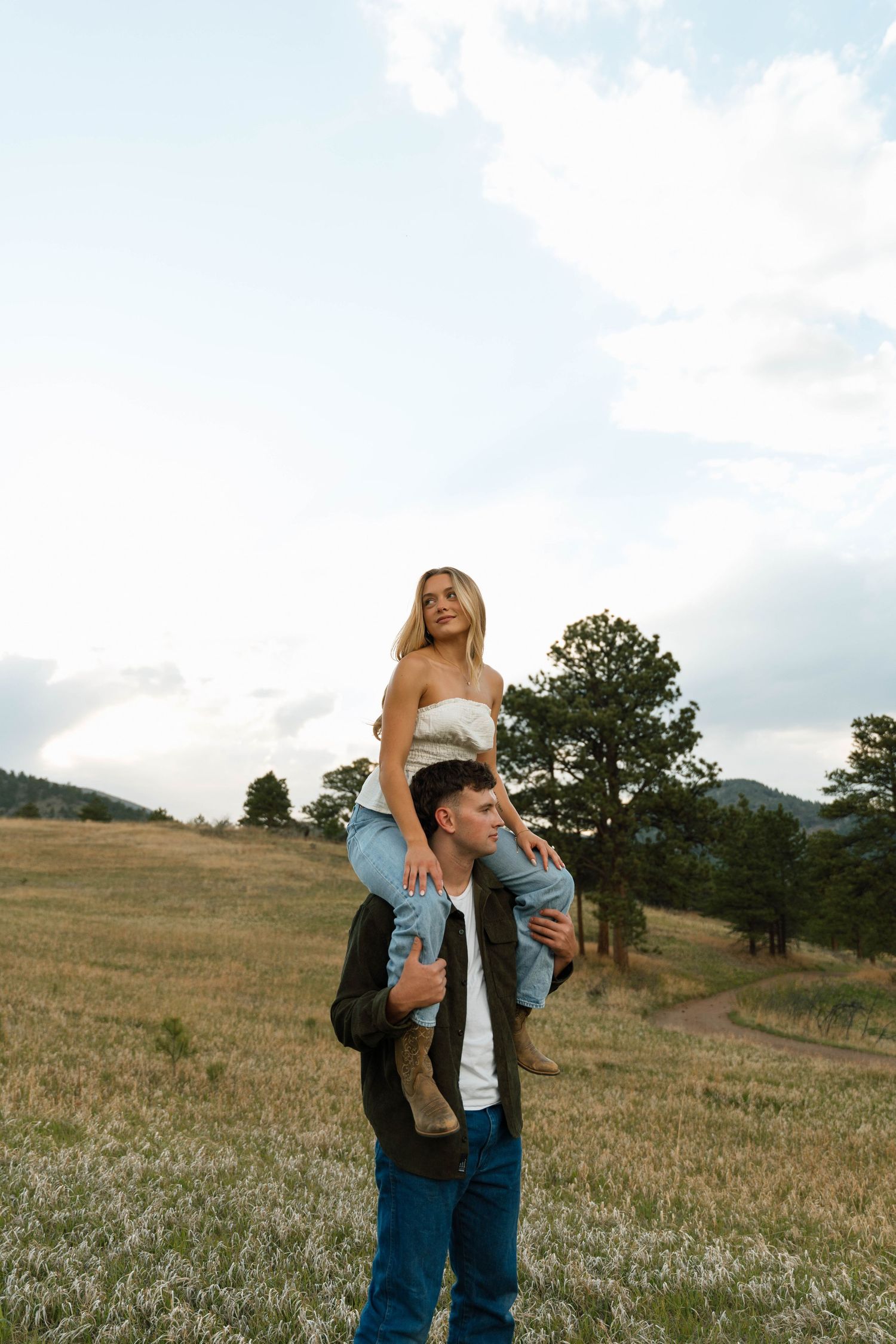 Man carrying woman on his shoulders in a scenic mountain meadow, both wearing jeans and casual tops.