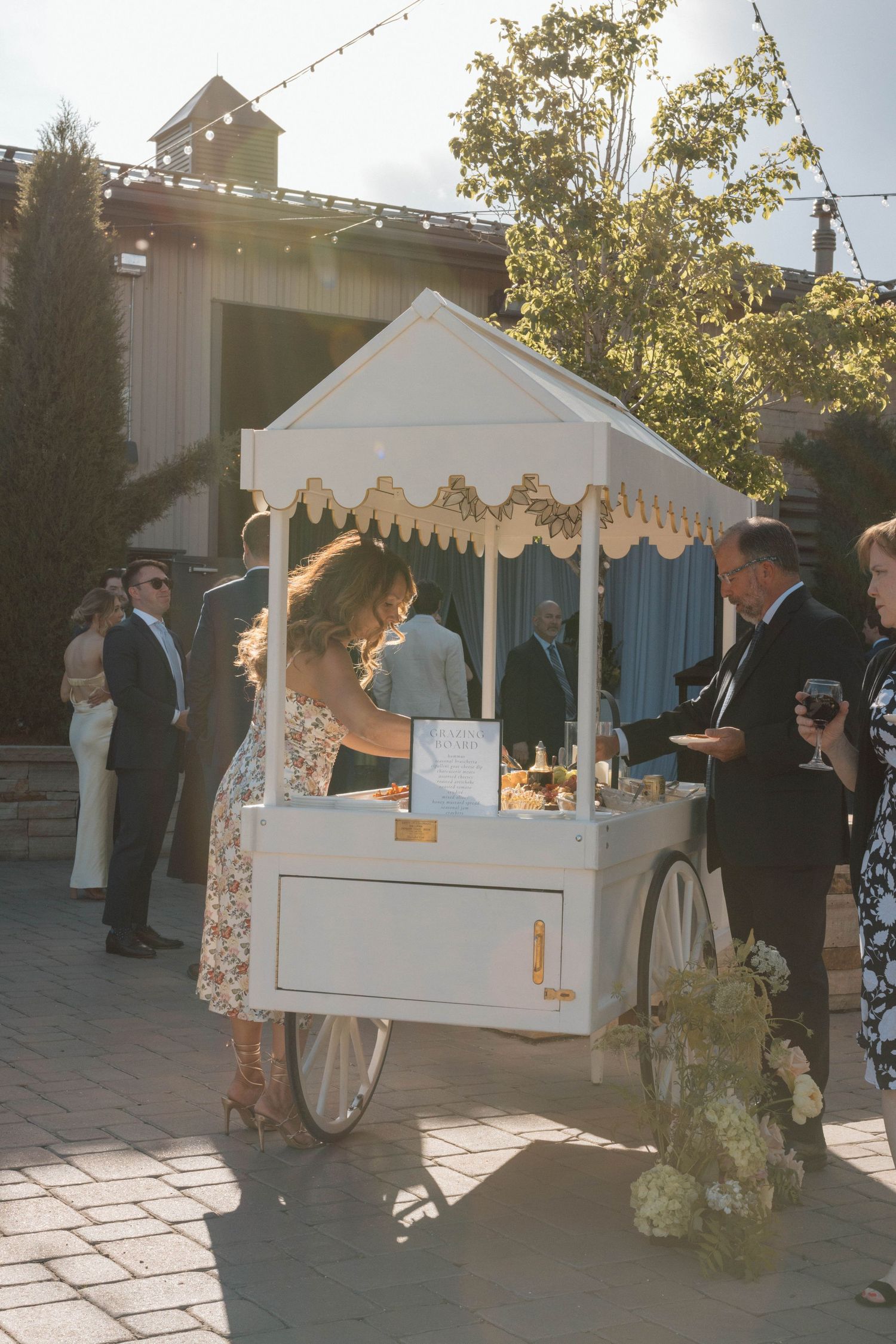 White vintage cart serving guests at outdoor wedding reception on sunny paved courtyard with string lights.