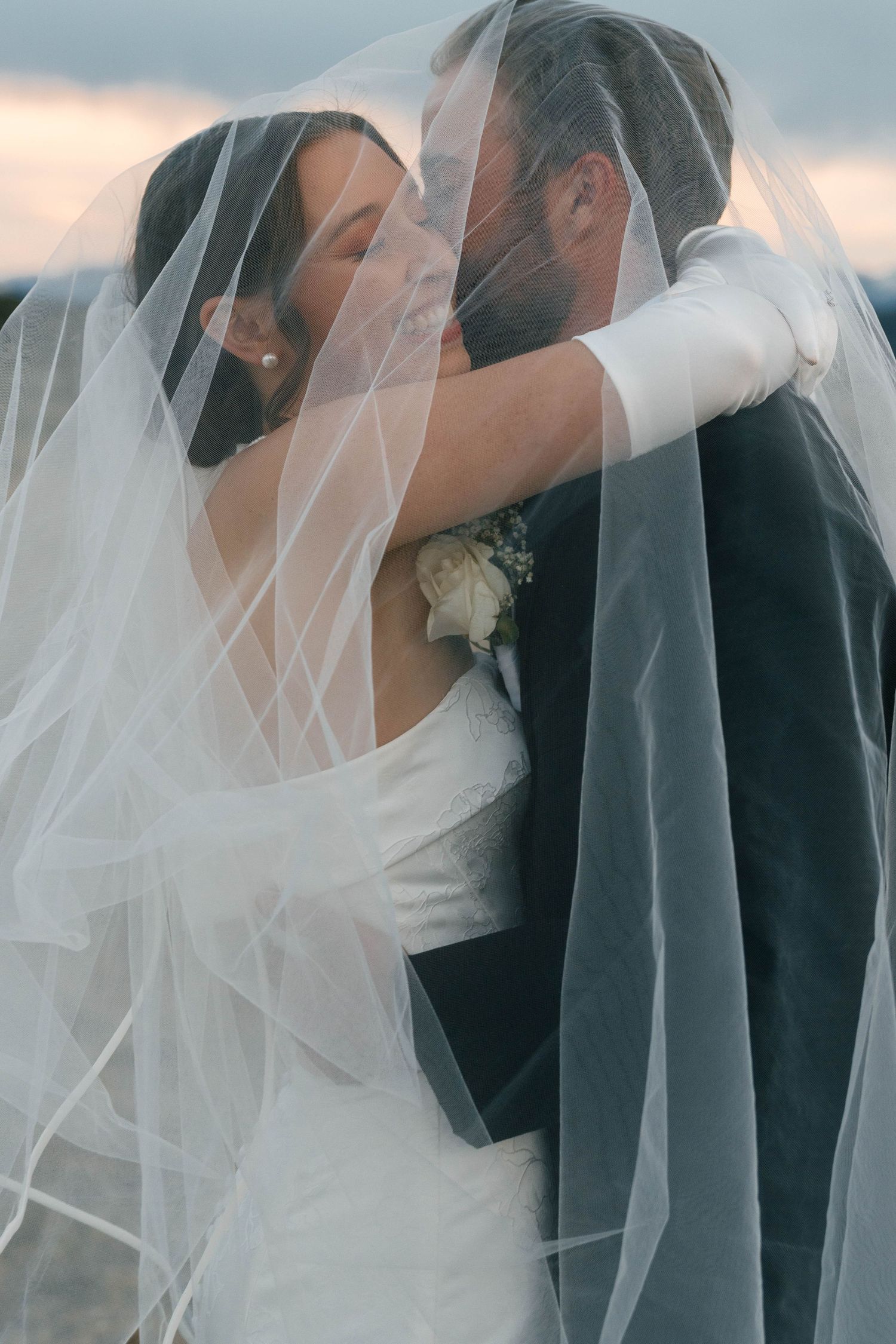 Bride and groom sharing a romantic kiss under a flowing white wedding veil at sunset.
