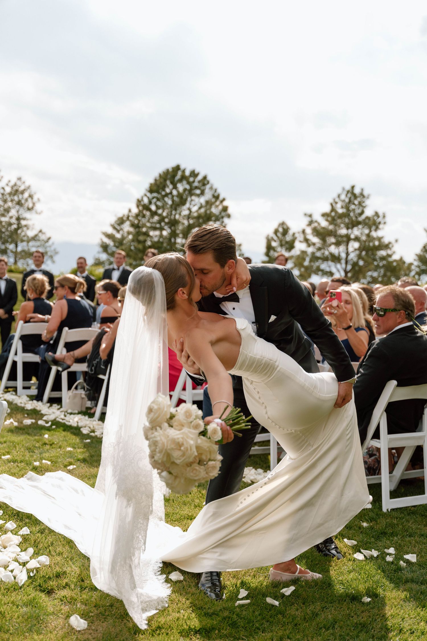 Romantic dip kiss during outdoor wedding ceremony with guests seated on white chairs.