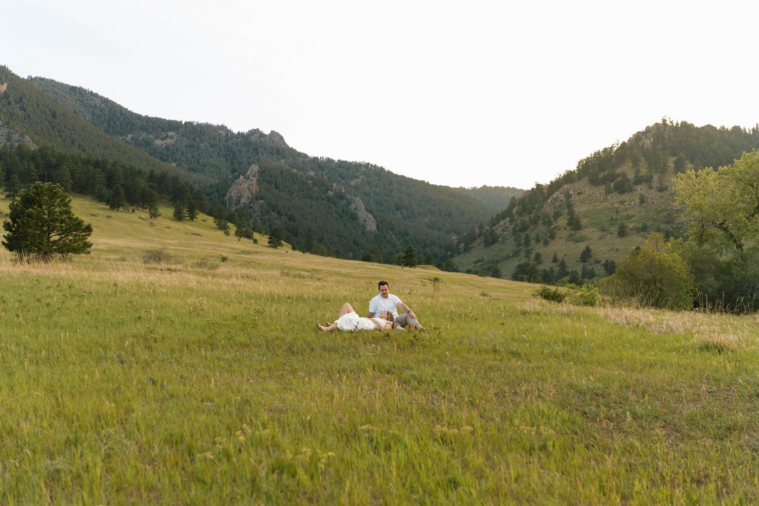 Couple sitting together in a lush green meadow surrounded by forested mountains during golden hour.
