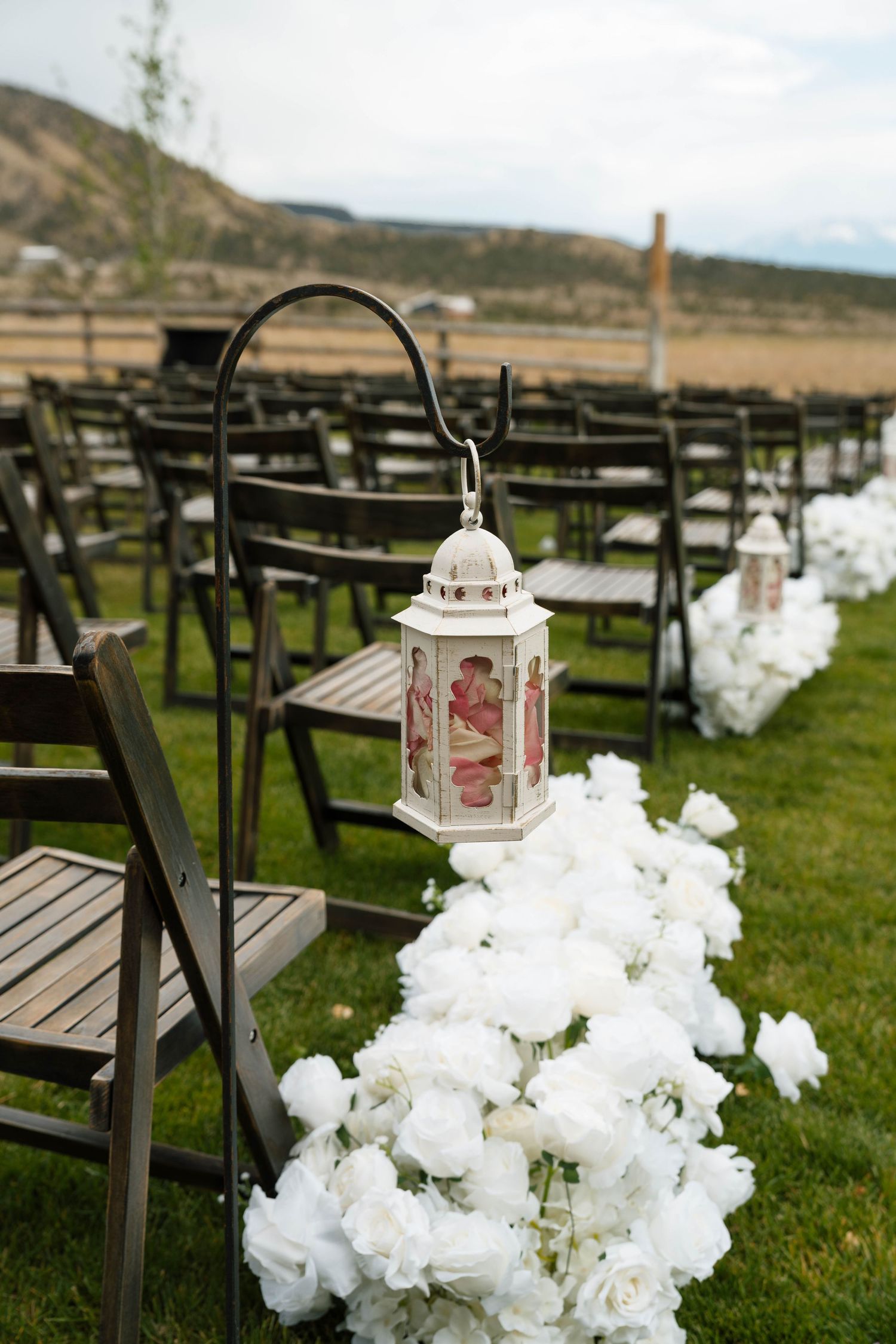 Elegant outdoor wedding aisle lined with white roses and decorative lanterns with wooden chairs.