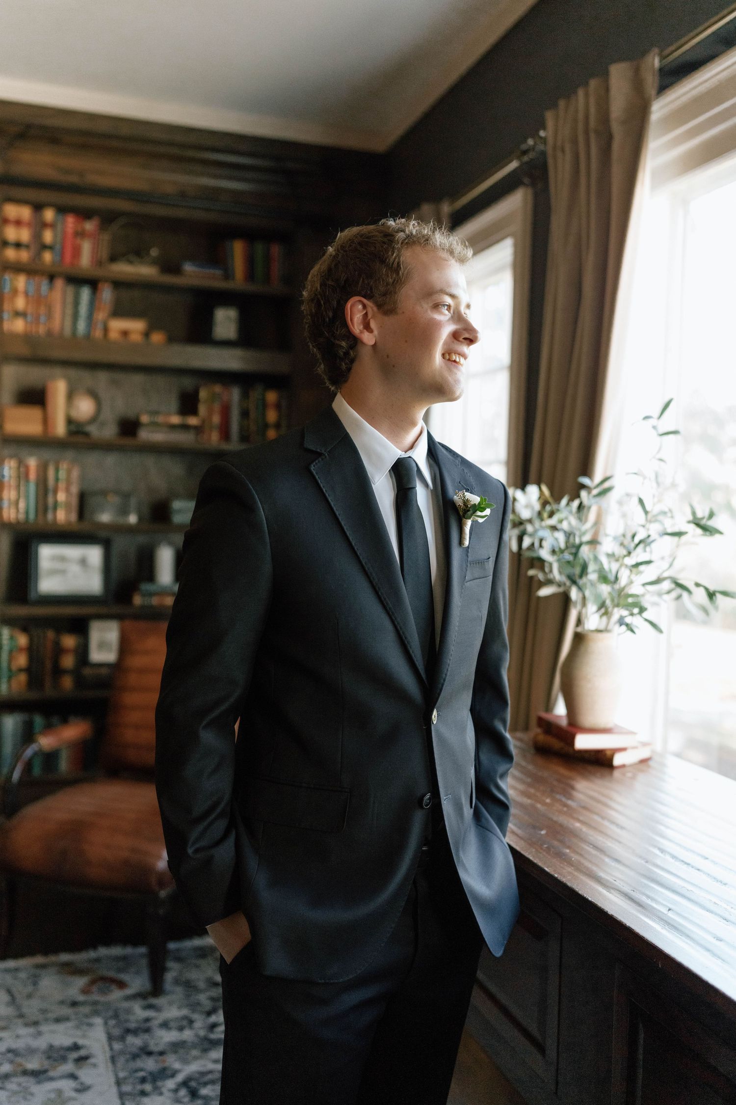 A person in a dark suit gazes thoughtfully out of a window in a study with bookshelves.