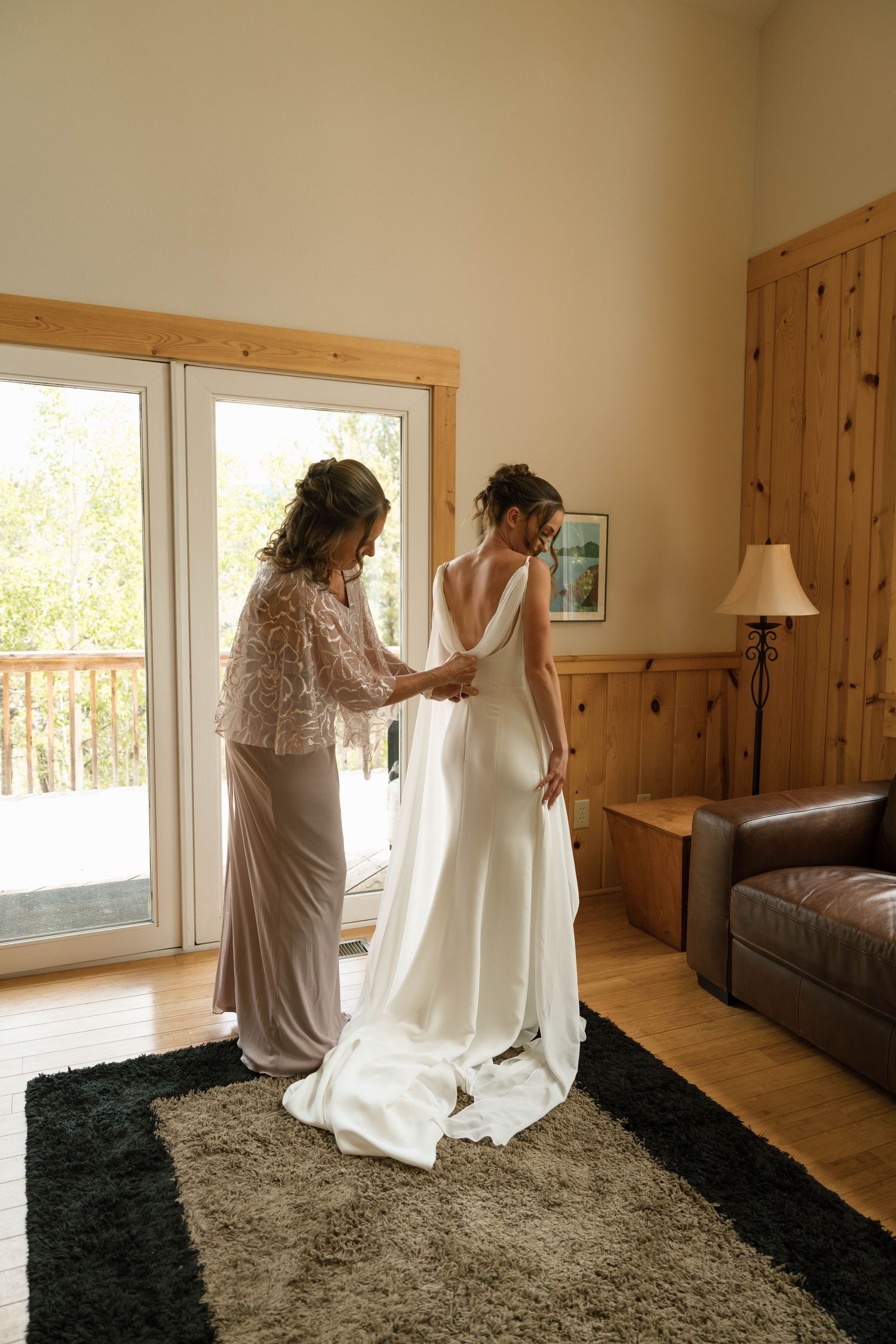A bride in a white gown gets her dress fastened by a woman in a lace gown inside a wood-paneled room.