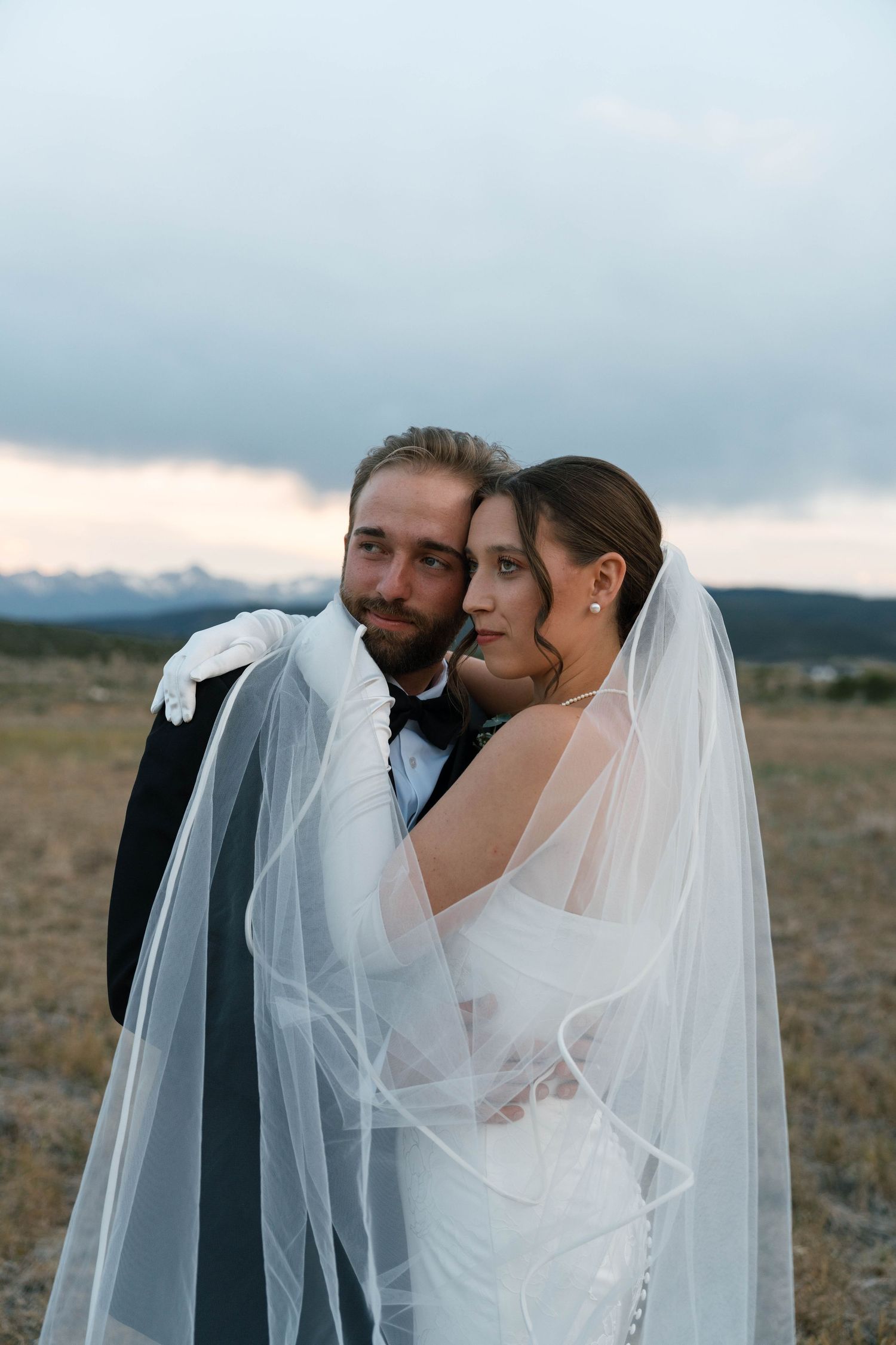 Bride and groom posing with flowing veil in rustic mountain field at twilight.