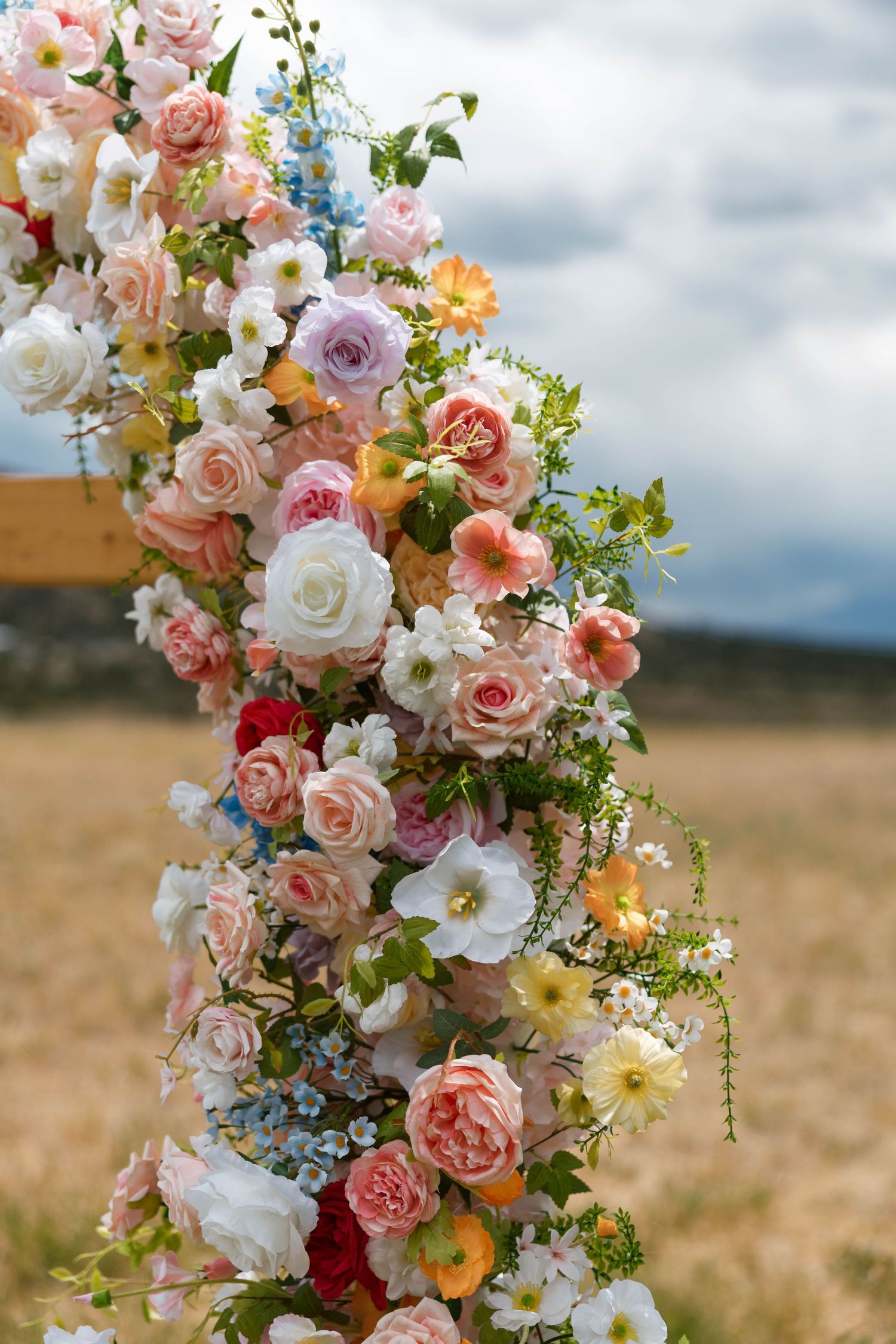 Colorful floral arch with pink, white, and blue roses for an outdoor wedding ceremony against a cloudy sky.