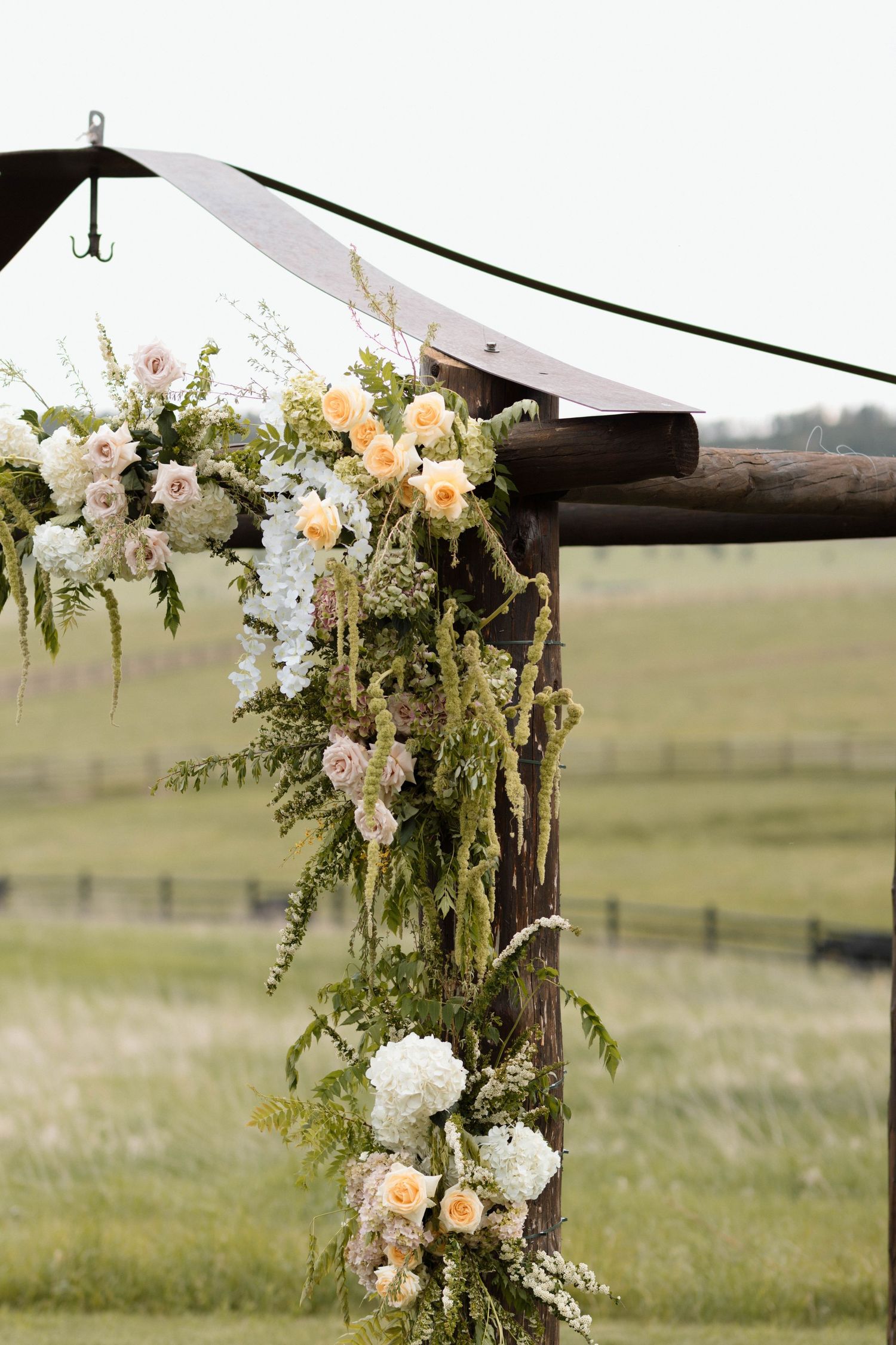 Rustic wooden wedding arch adorned with peach roses, white hydrangeas, and cascading greenery in an outdoor field setting.