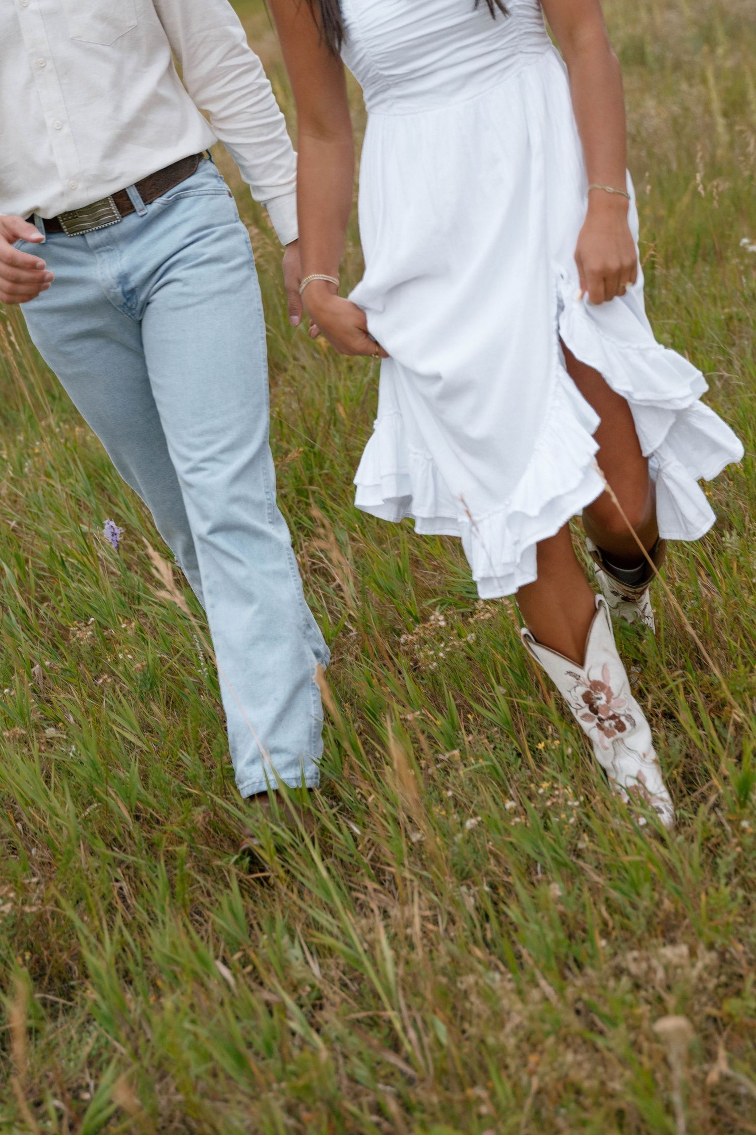 Close-up view of a couple's legs walking through tall grass, wearing white dress and jeans.
