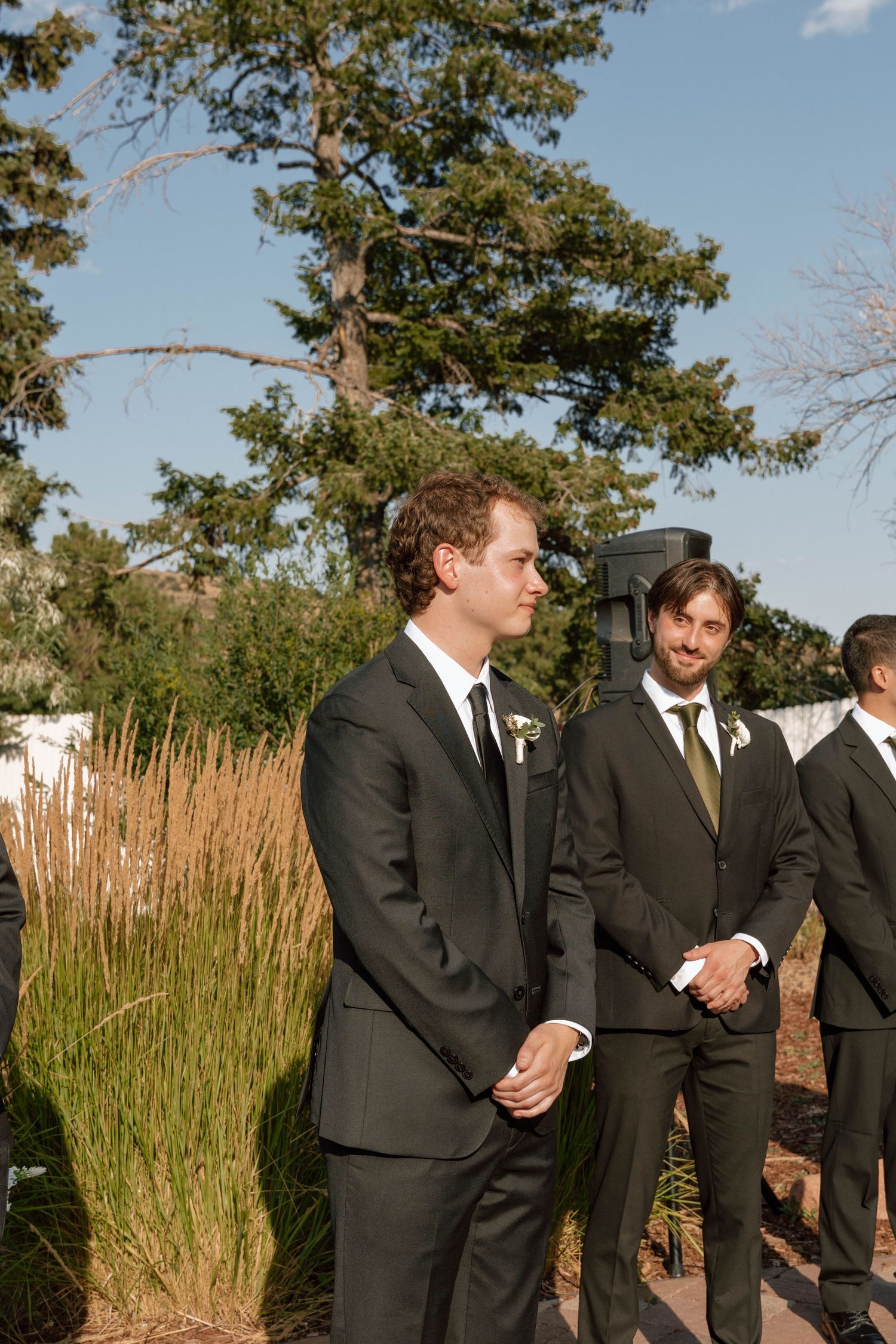 Groomsmen in dark suits stand together during an outdoor wedding ceremony.