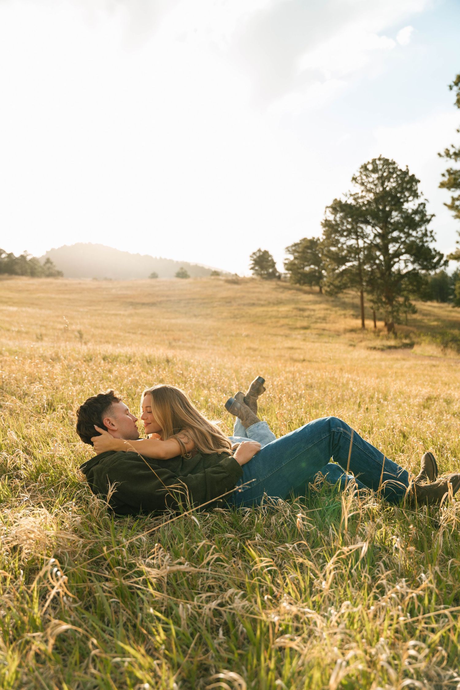 Couple lying in a sunlit golden meadow, sharing a romantic moment with pine trees and mountains in the background.