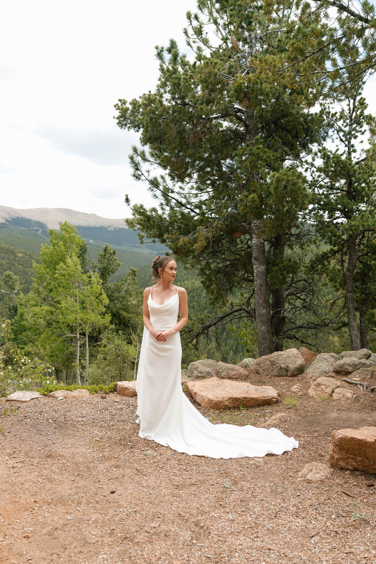Bride in white gown stands on rocky mountain overlook surrounded by pine trees and scenic Colorado landscape.