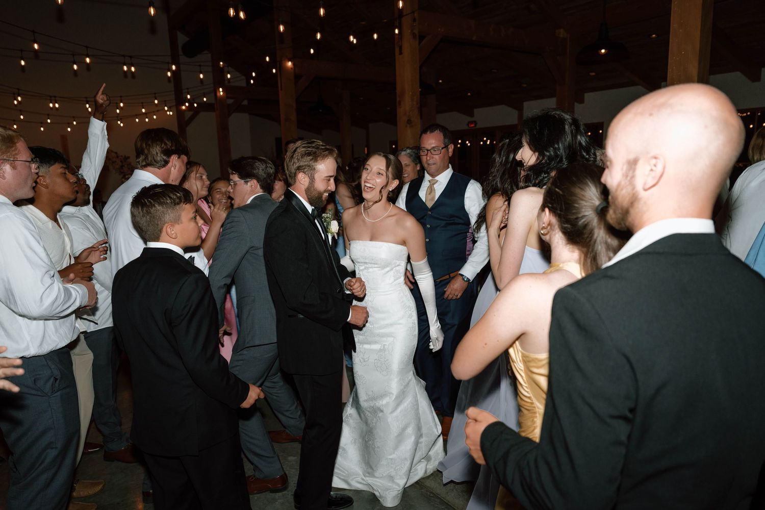 Bride and groom celebrate with guests on the dance floor at lively indoor wedding reception.