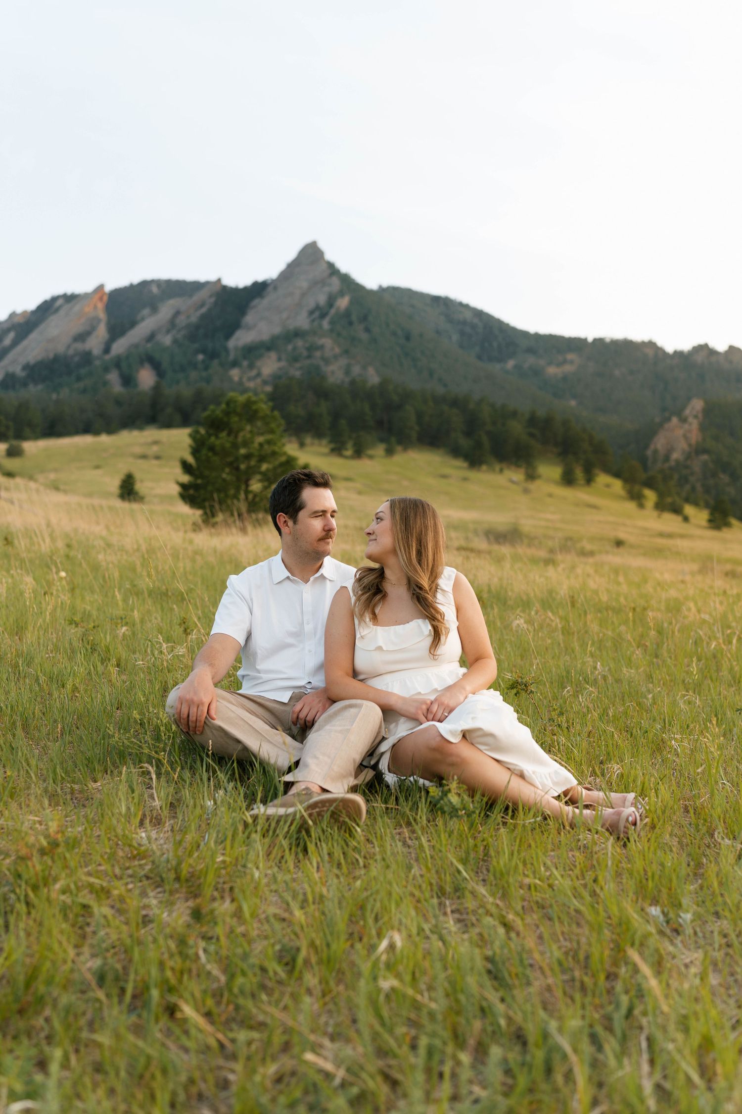 Couple sitting together in a green meadow with rocky mountain flatirons in the background on a sunny day.