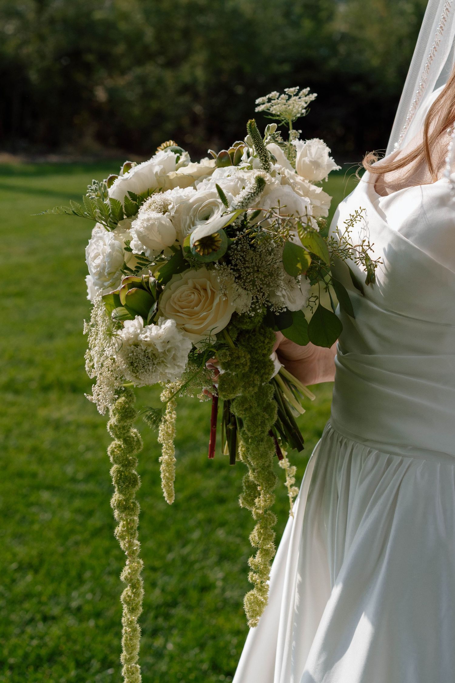 White floral bouquet with cascading greenery held against a green outdoor background.