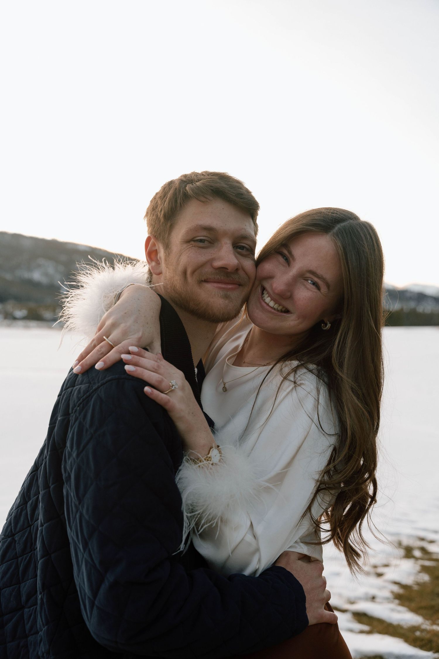 Joyful couple sharing laughter during their winter engagement session by a lake.