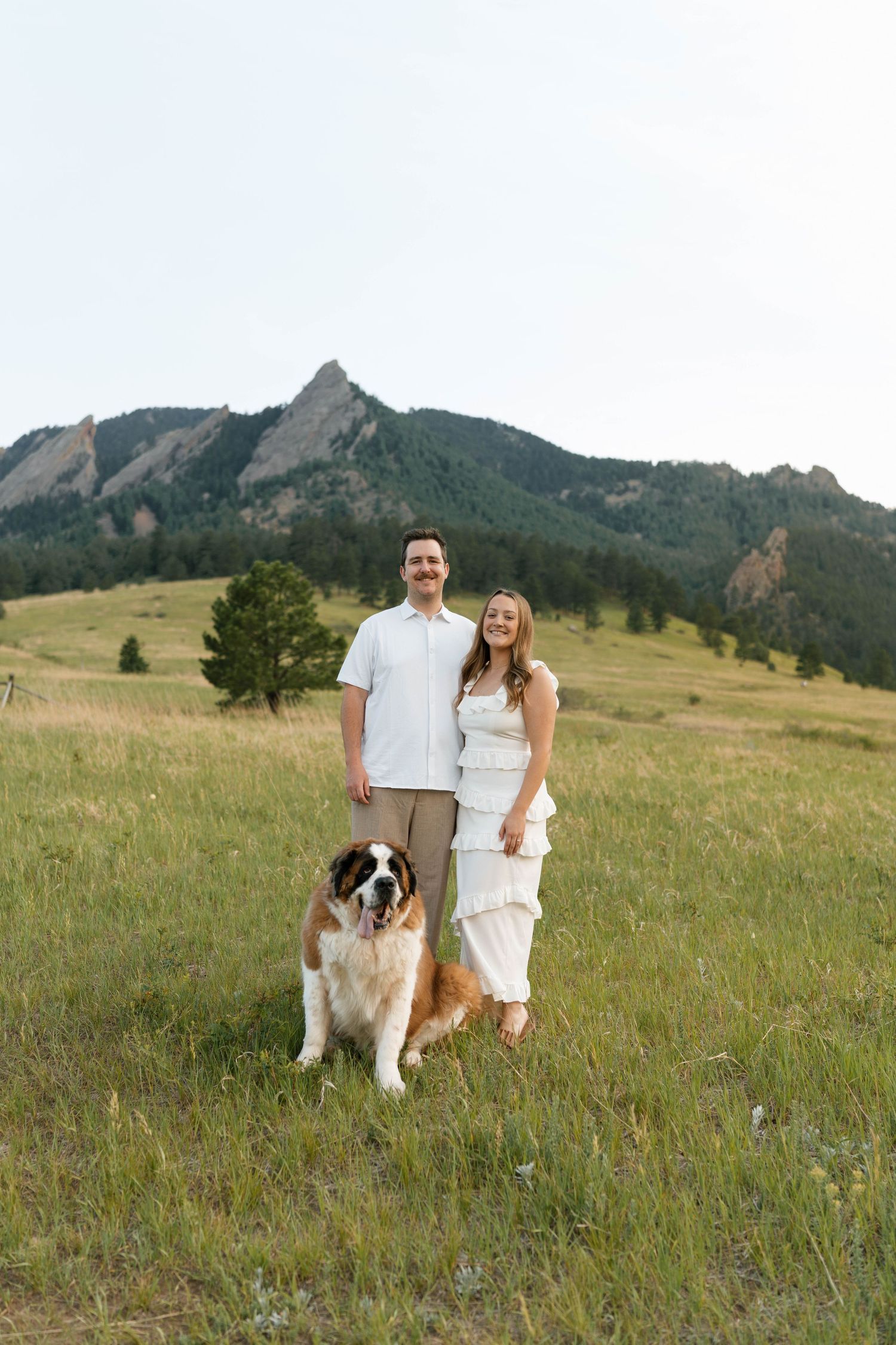 Couple smiling with Saint Bernard dog in green meadow with rocky Flatirons mountains in background.