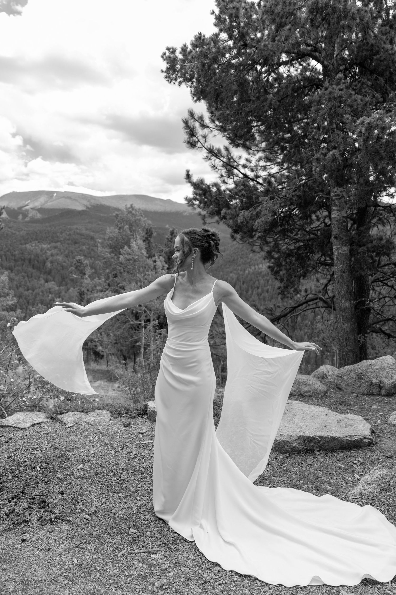 Bride in flowing white wedding gown with dramatic cape sleeves poses on rocky mountain overlook.