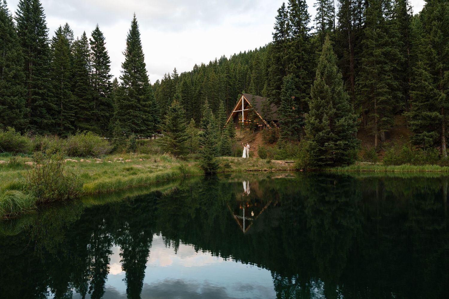 A rustic cabin nestled among tall pine trees reflecting on a calm mountain lake at dusk.