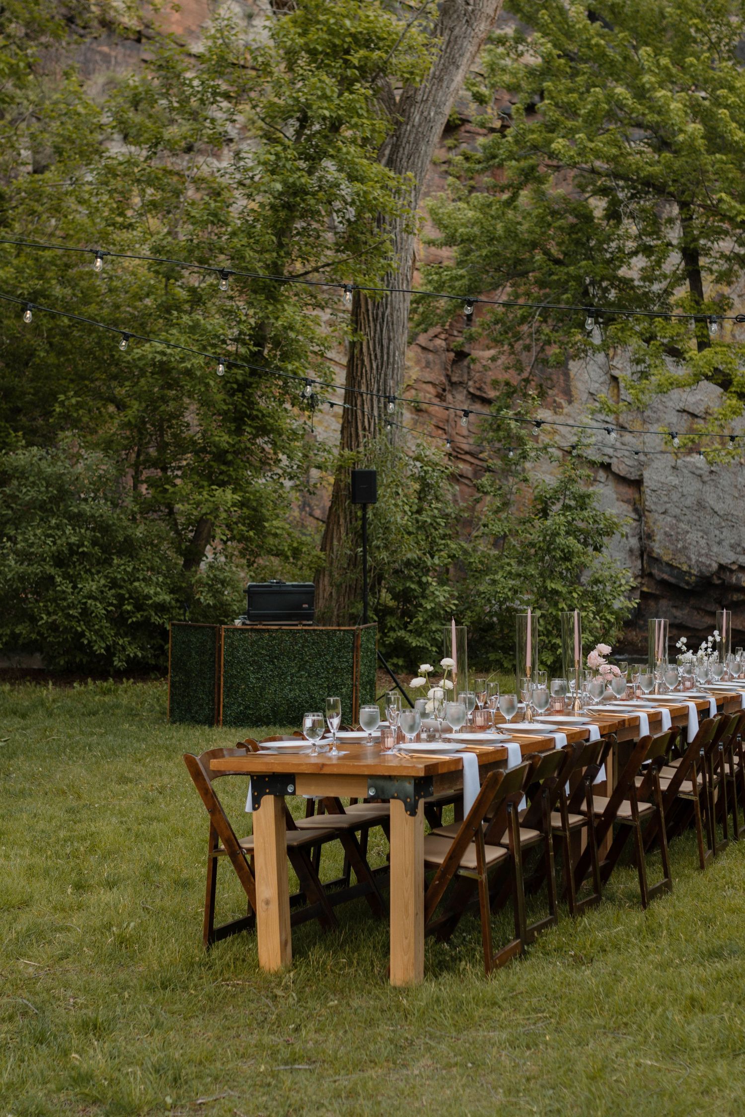 Elegant outdoor wedding reception table set on grass with wooden chairs, floral centerpieces, and rocky cliffside backdrop.