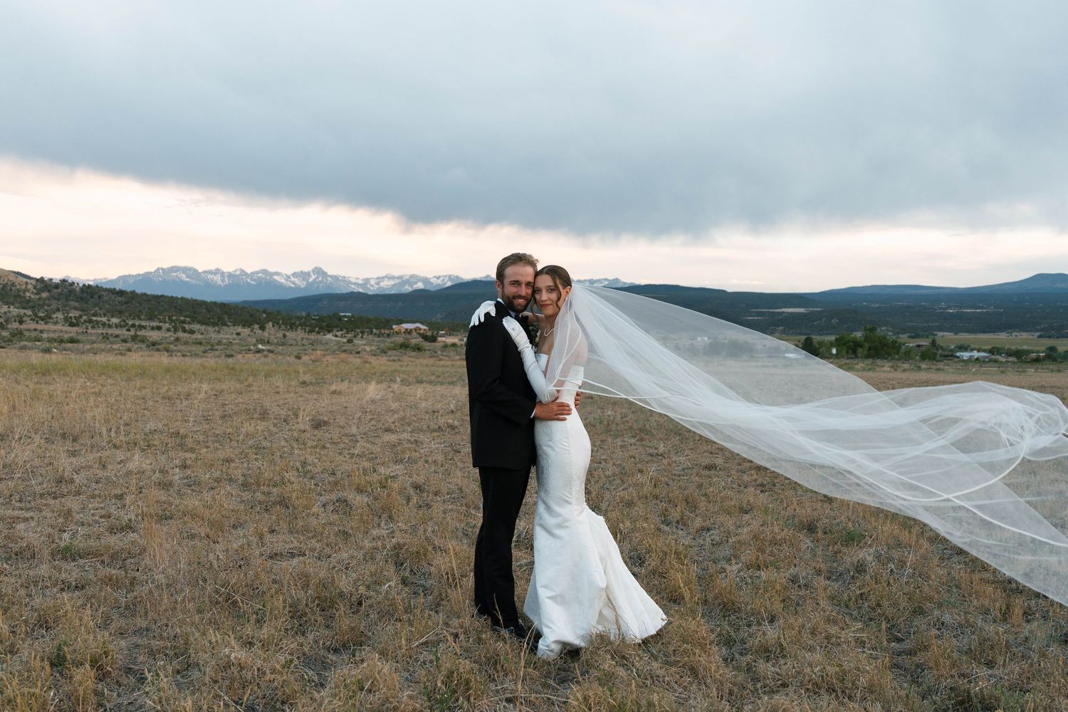 Bride and groom embrace in a mountain meadow, her cathedral veil flowing in the wind under a dramatic cloudy sky.