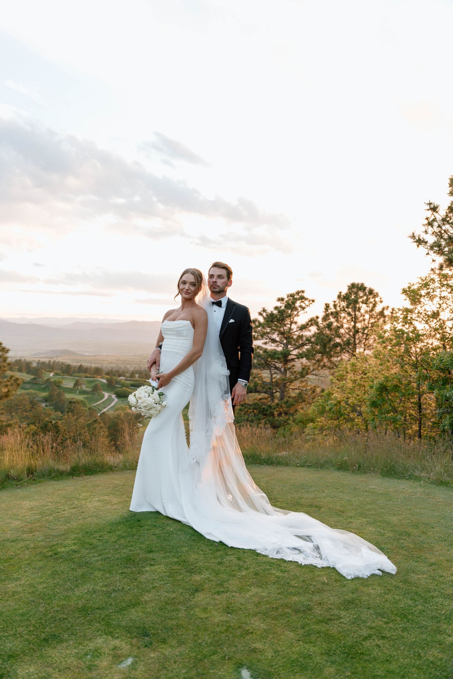 A couple shares an intimate moment at sunset on a hilltop with a flowing white wedding dress trailing behind them.
