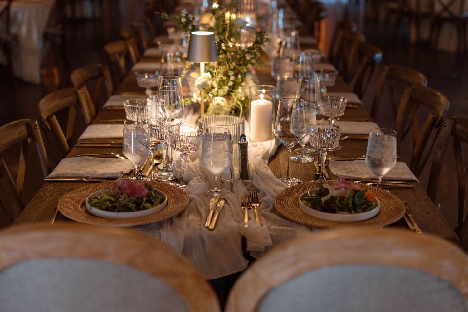 Elegant wedding reception table set with gold chargers, crystal glasses, candles, and lush greenery centerpieces.