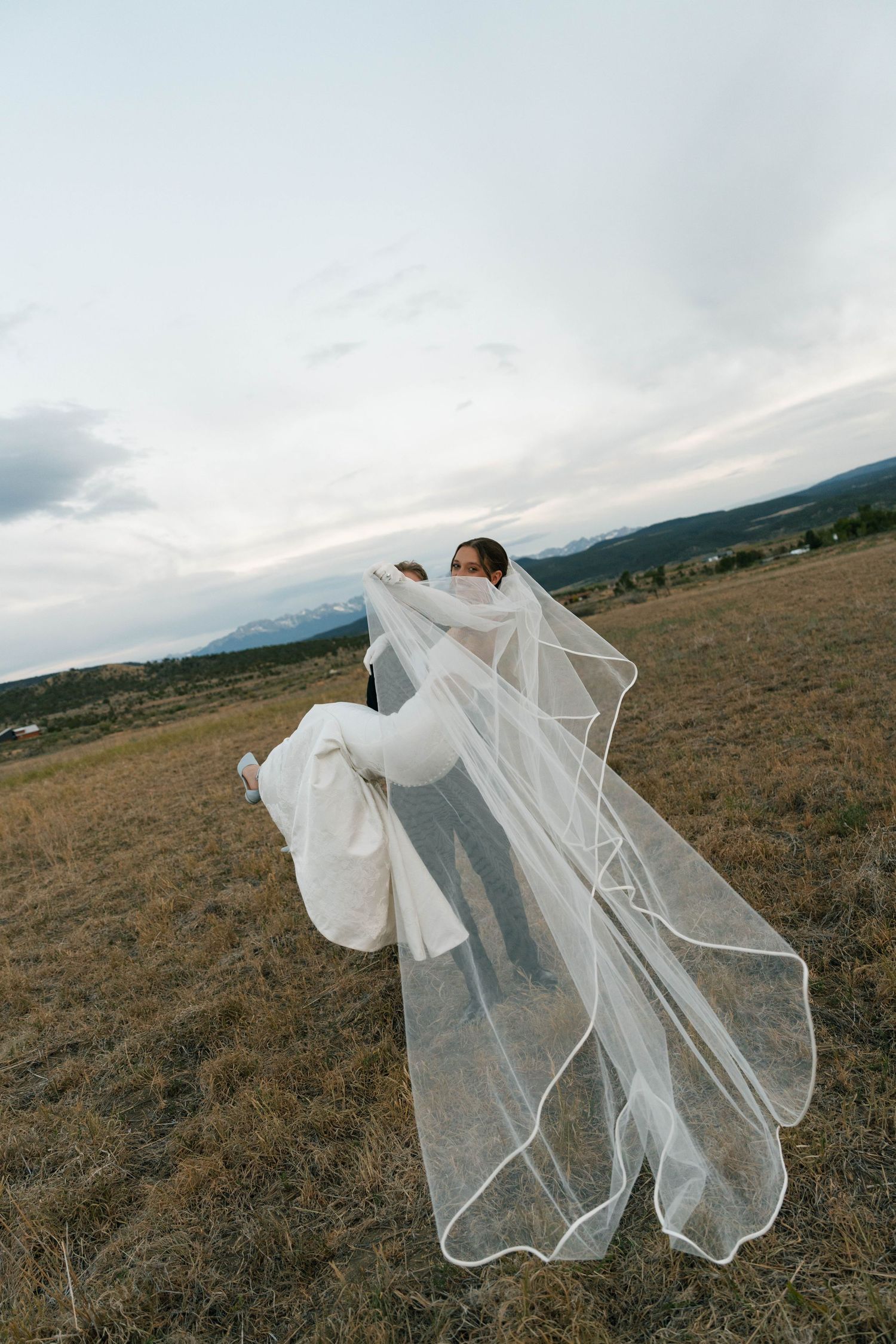 A bride in a flowing white gown and veil being lifted by groom in an open field with mountains in the background.