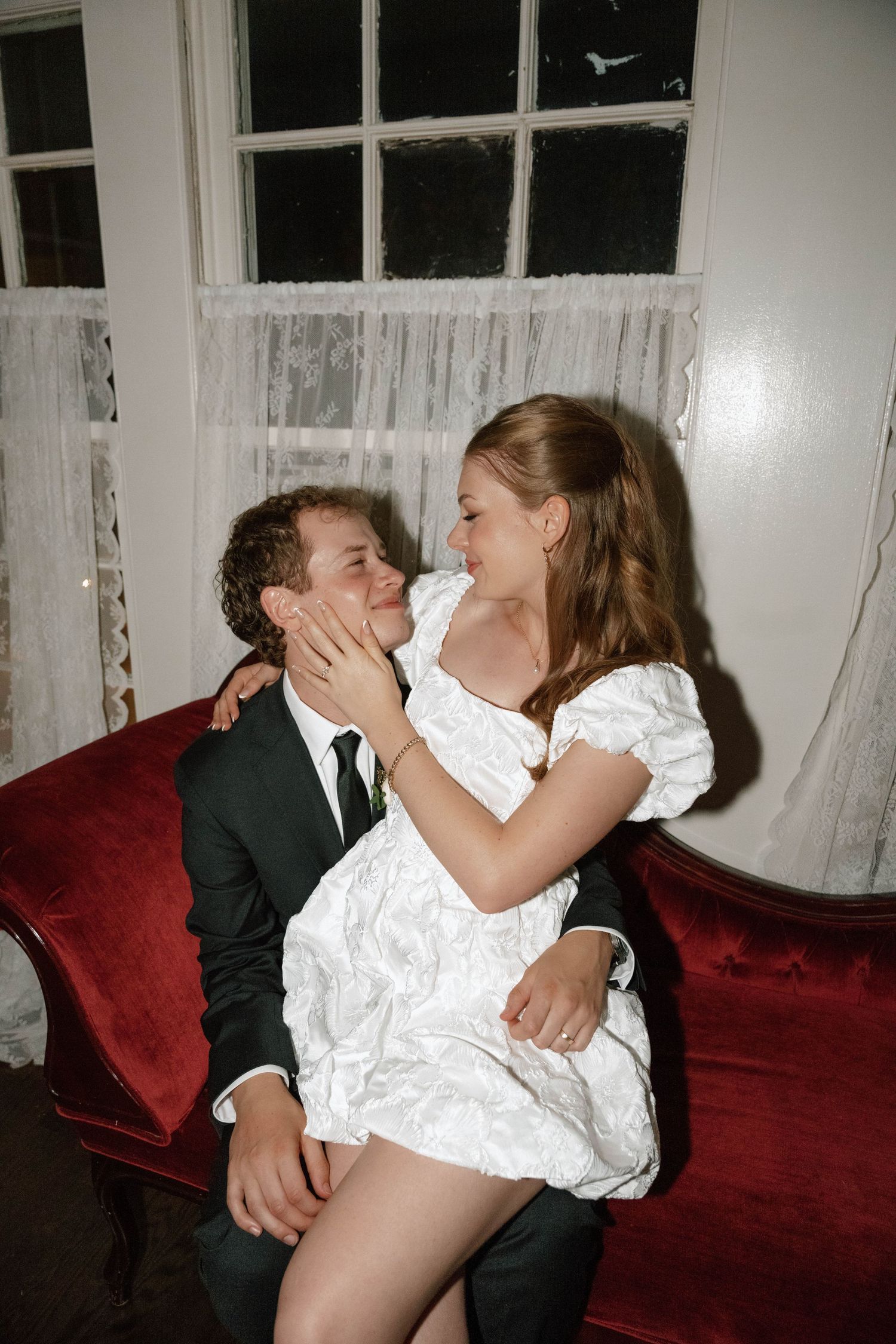 An intimate moment shared between a couple on a red velvet sofa in a vintage-style setting.