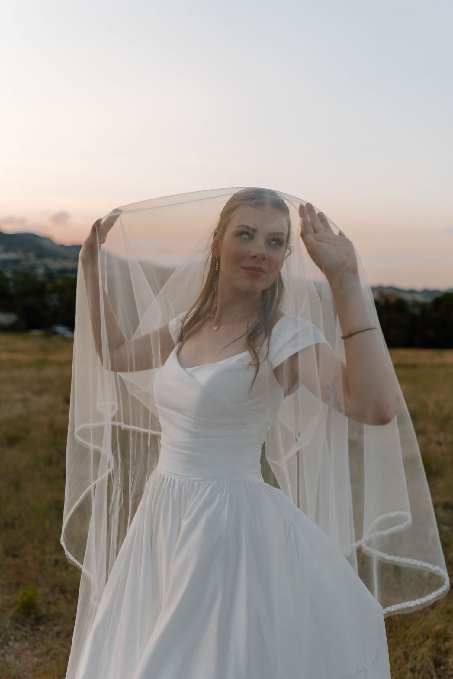 Bride adjusts flowing veil in mountain field during golden hour.