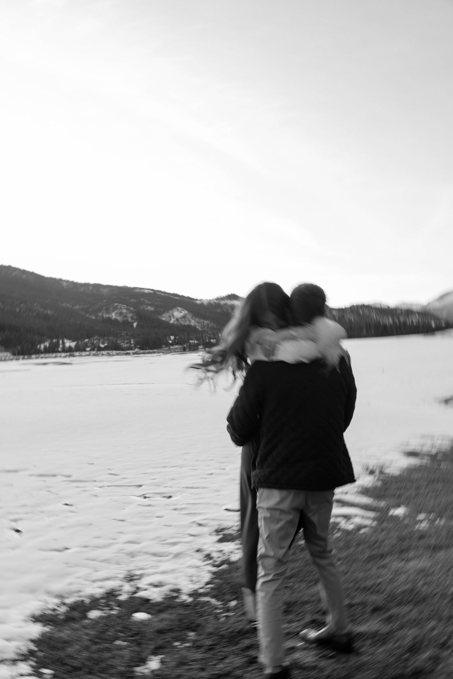 A romantic couple embraces against a snowy mountain landscape in black and white.