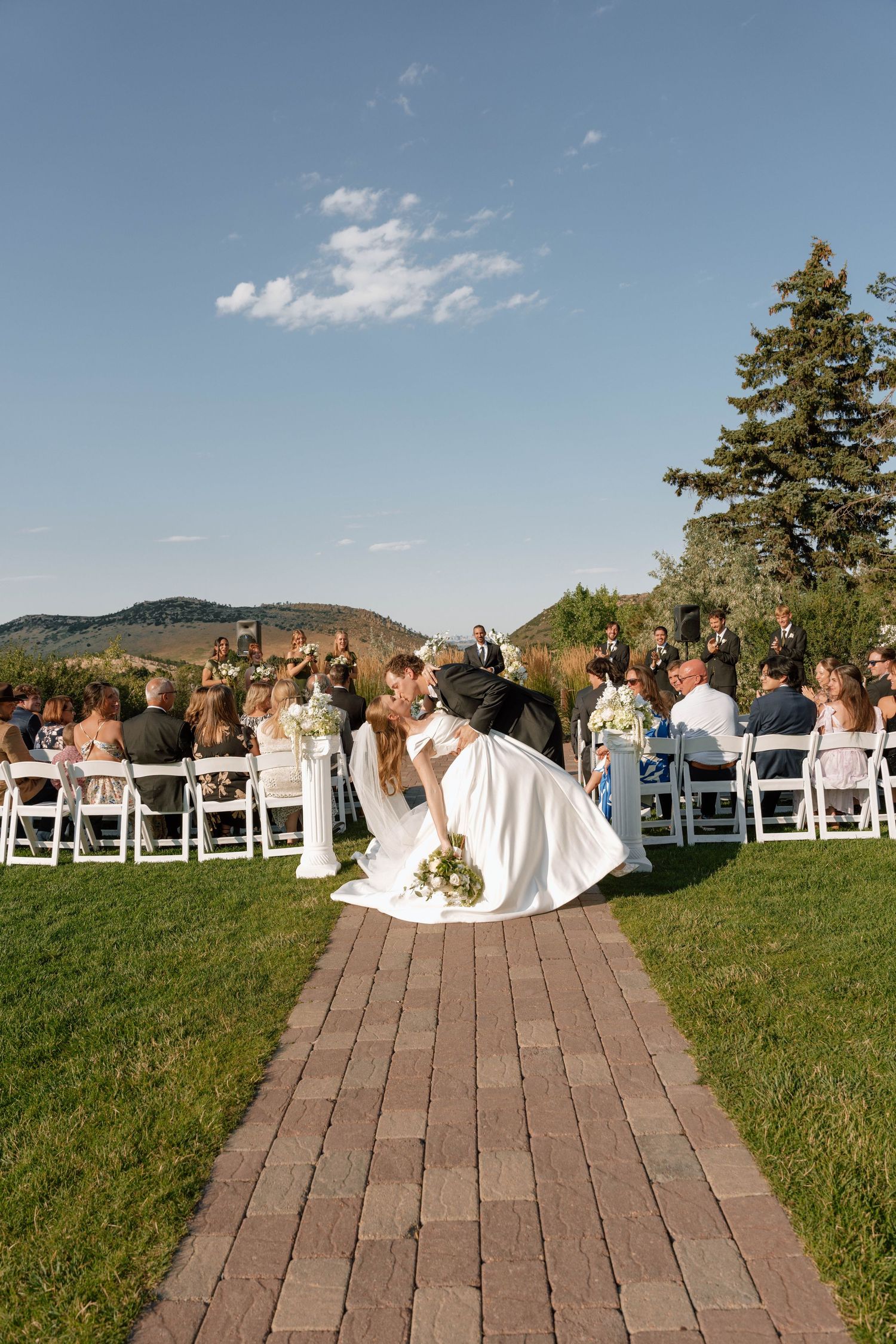 Bride and groom share a dip kiss at outdoor wedding ceremony on brick path with guests seated on white chairs.