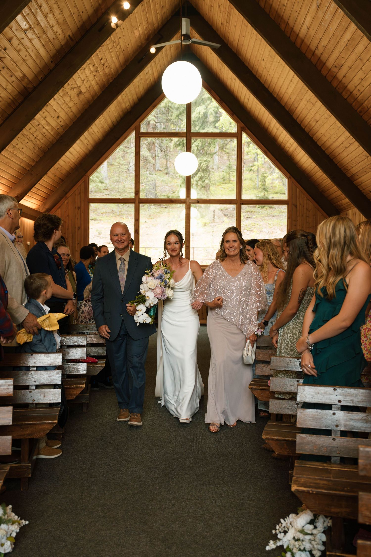 Bride walking down chapel aisle escorted by parents in rustic wood A-frame venue.