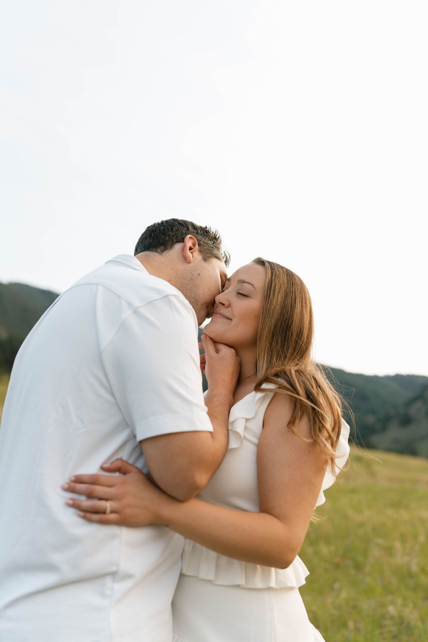 Man kissing womans cheek while embracing in green mountain meadow on a sunny day.