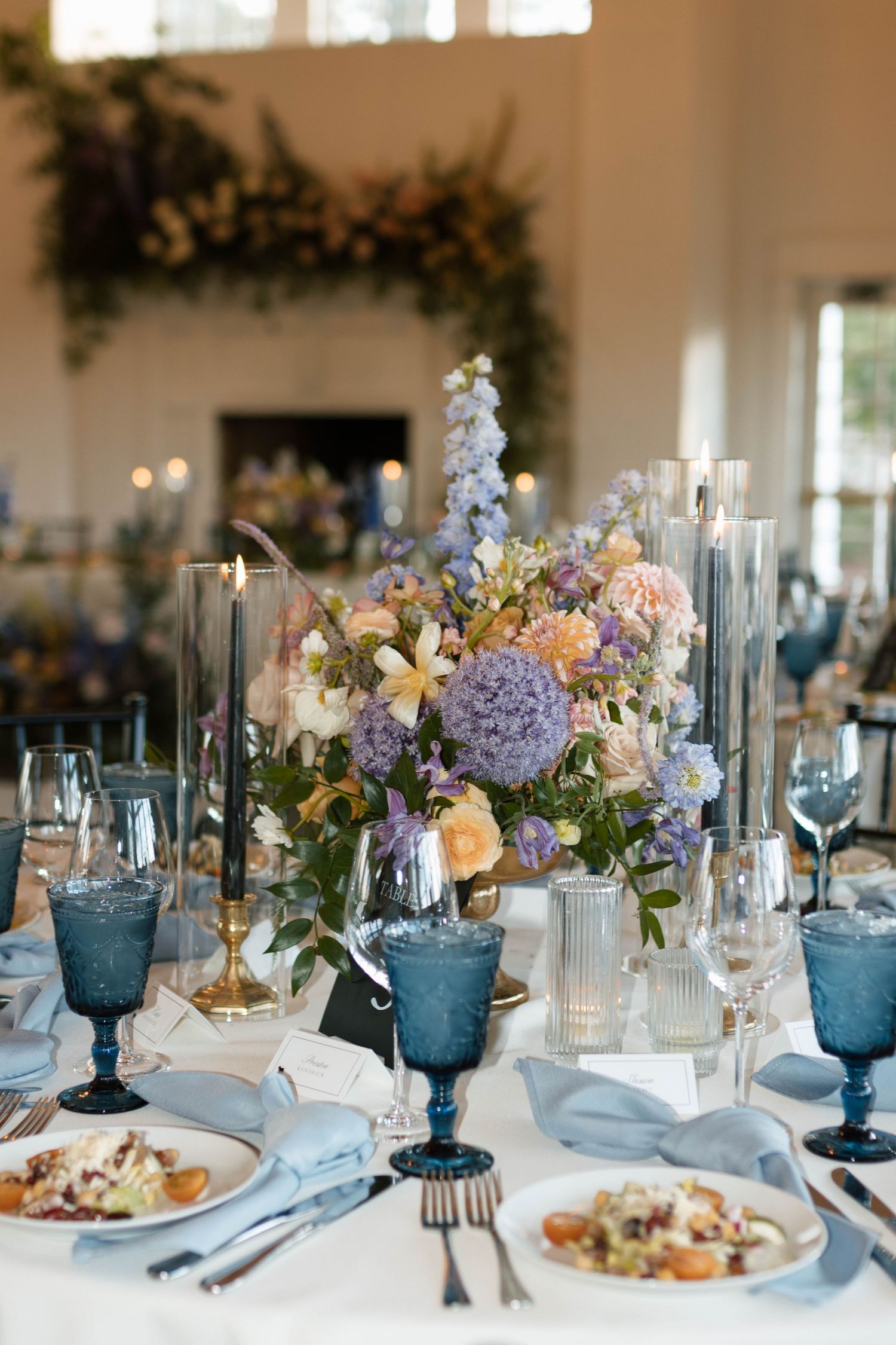 Elegant wedding table setting with blue glassware, white linens, and a lush floral centerpiece of hydrangeas and roses.