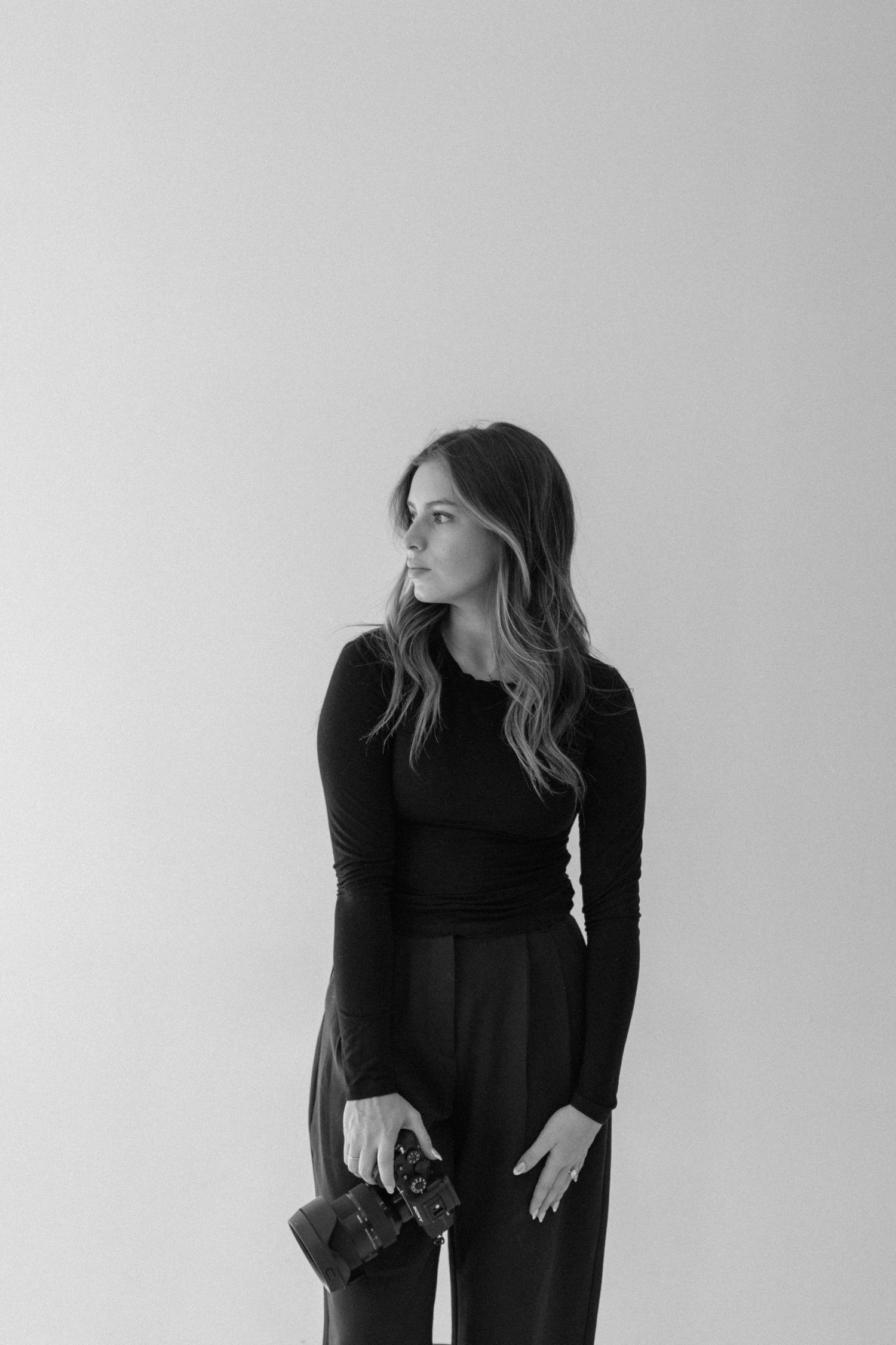 Black and white portrait of a woman in black outfit holding a camera, looking away against a light background.
