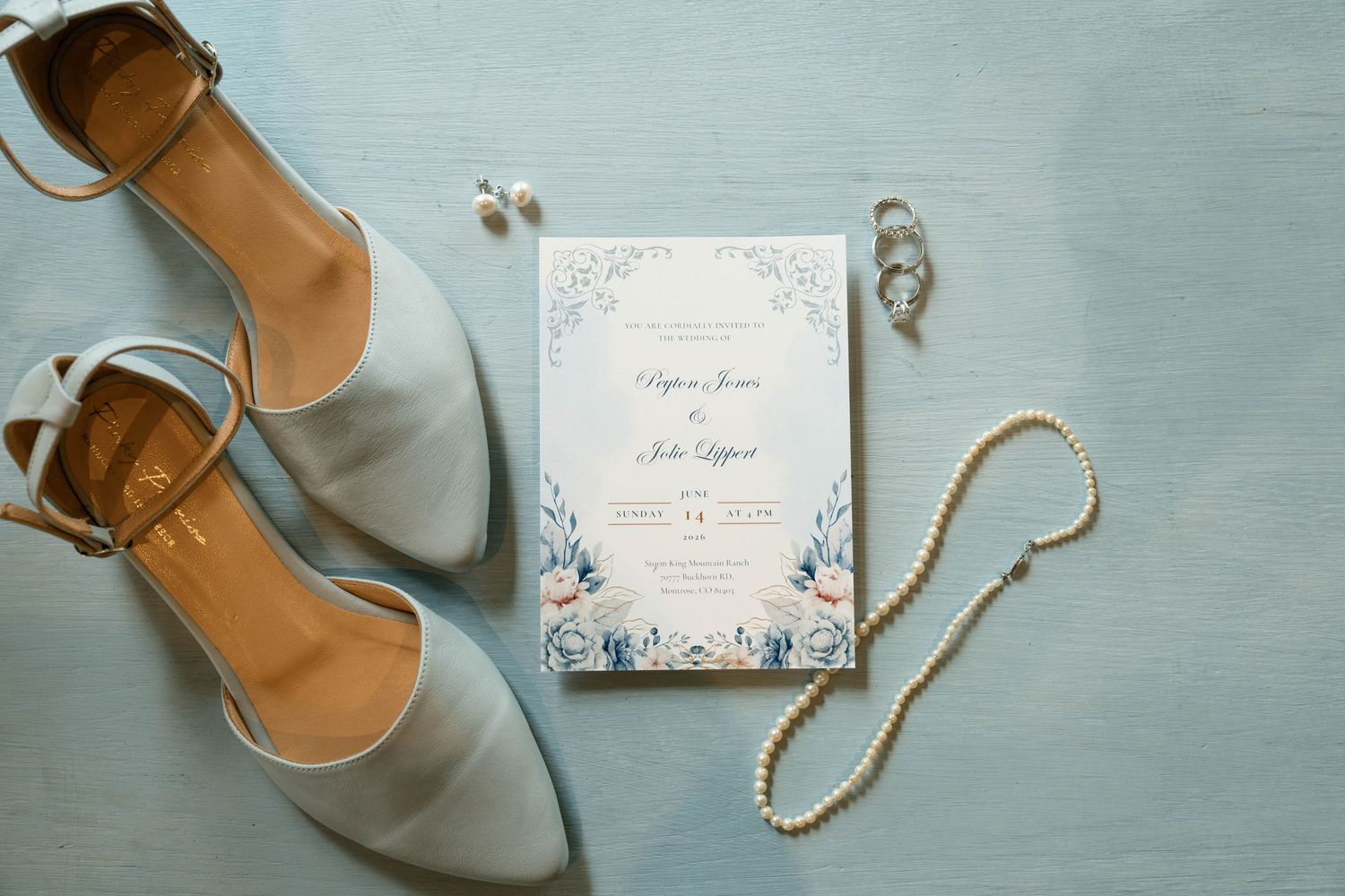 Wedding flat lay with blue heels, floral invitation, pearl necklace, and rings on a light blue background.