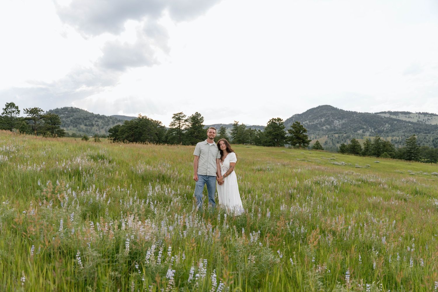Couple standing in wildflower meadow with mountain landscape behind them.